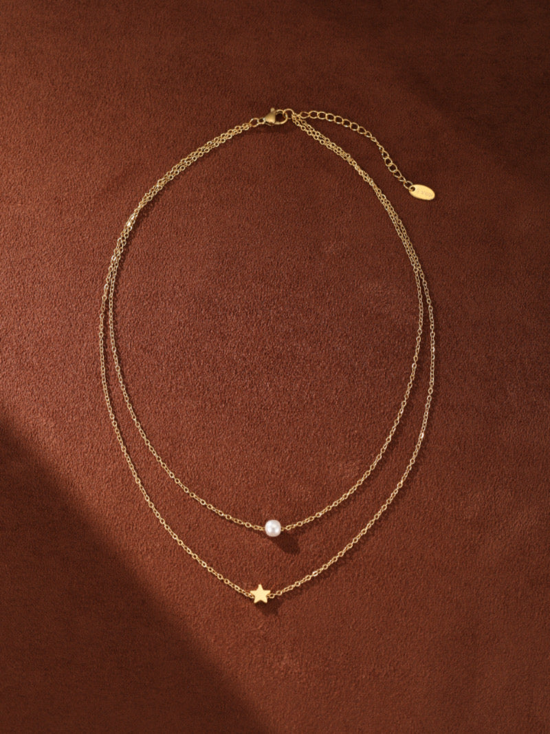 Un collier superposé ton or présentant une délicate chaîne avec une petite perle blanche d'eau douce et une seconde chaîne avec une breloque étoile, sur fond texturé marron.