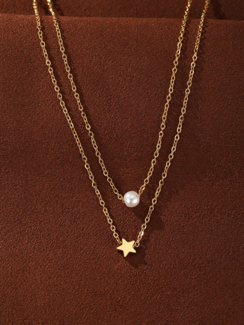 Collier superposé ton or avec une seule perle blanche d'eau douce sur la chaîne supérieure et une petite breloque étoile sur la chaîne inférieure, contre un fond texturé marron.