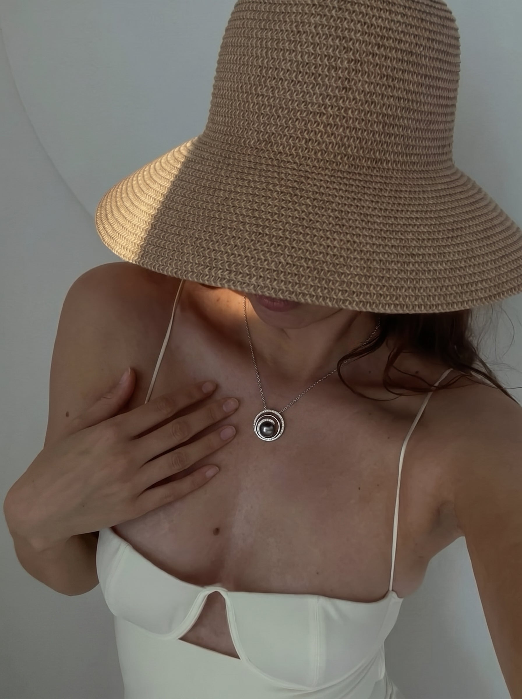 Une femme portant un chapeau de paille et une robe blanche, avec le collier pendentif concentrique Lunar Core en perle de Tahiti visible sur sa poitrine. Le collier présente une sombre perle de Tahiti entourée d'anneaux concentriques en argent sterling. @lifestyle