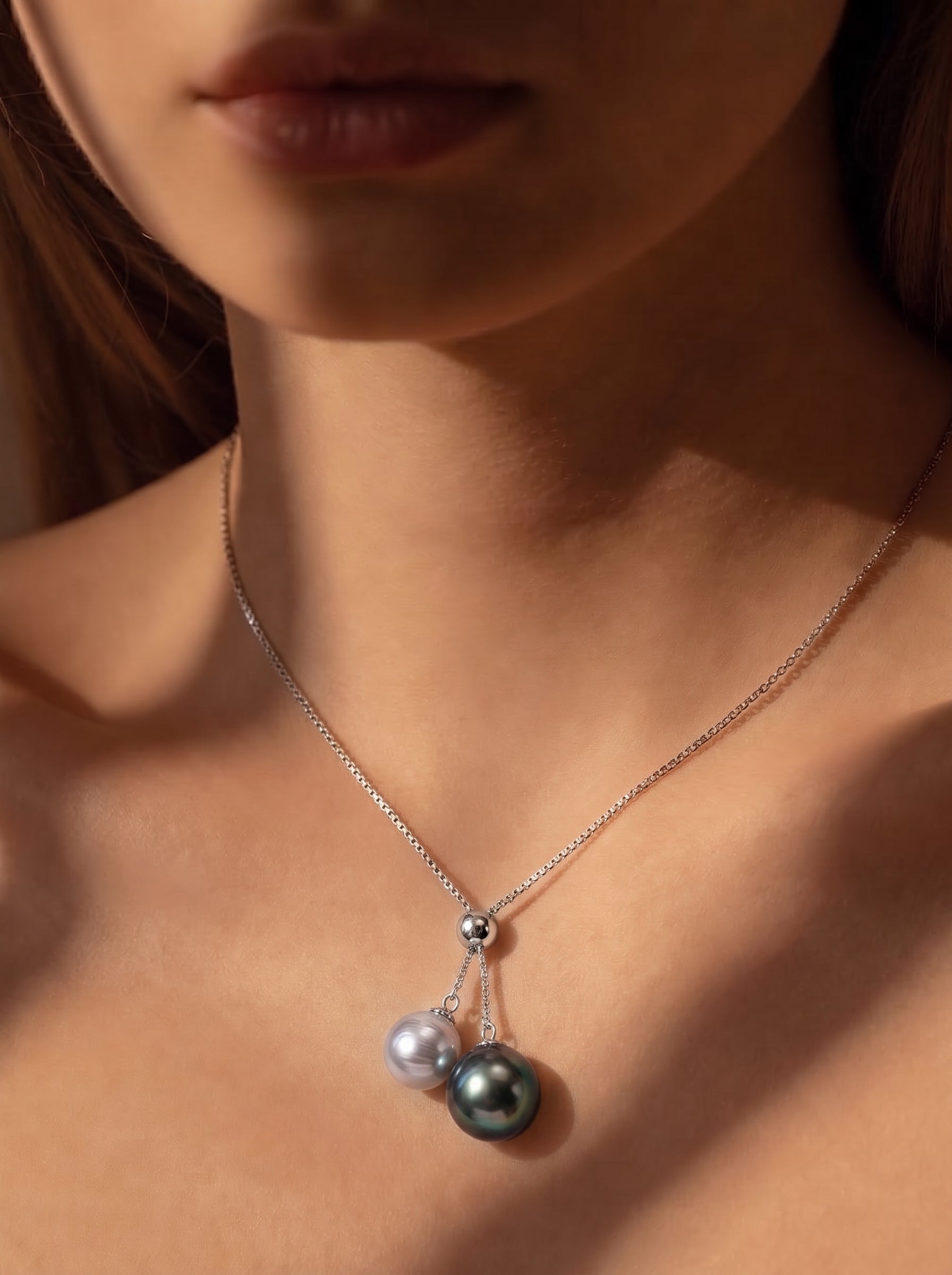 A close-up shot of a model wearing a delicate silver chain necklace with a Y-drop pendant featuring a Tahitian pearl and an Akoya pearl. @model