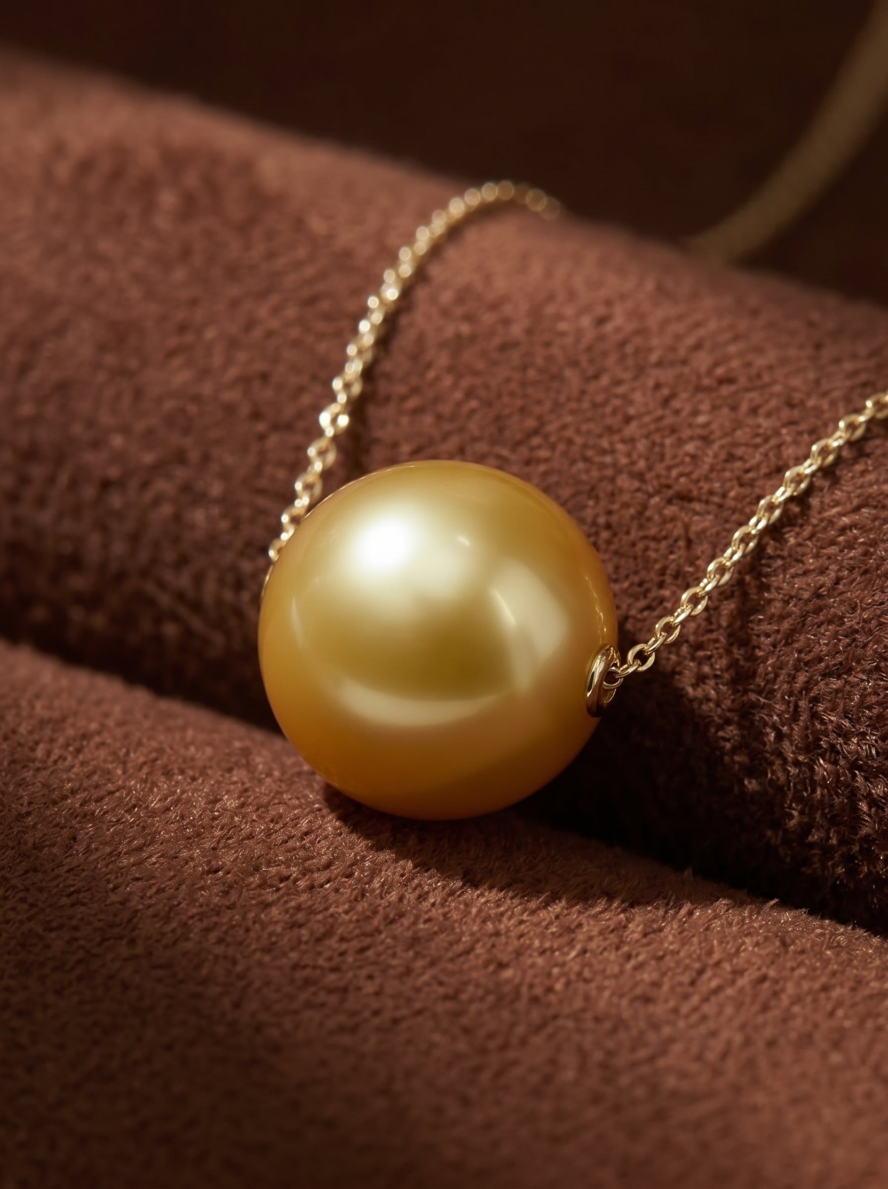 A close-up shot of a 12mm round Golden South Sea Pearl with exceptional luster, floating on a delicate gold chain. The pearl is showcased against a soft brown textured background. @pearl