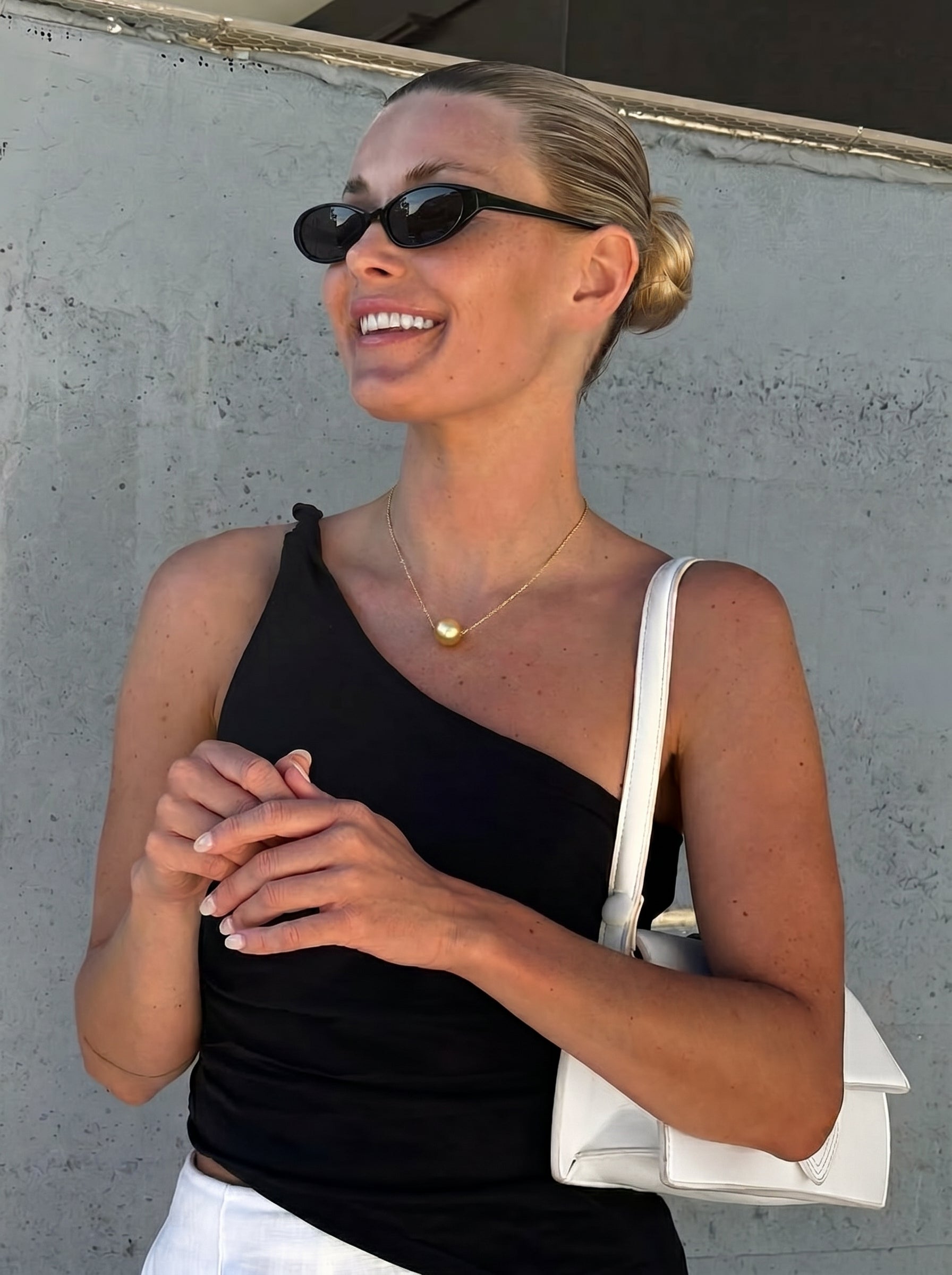 A smiling blonde woman in sunglasses and a black one-shoulder top wears a luminous Luster South Sea pearl floating necklace against a concrete wall. @lifestyle