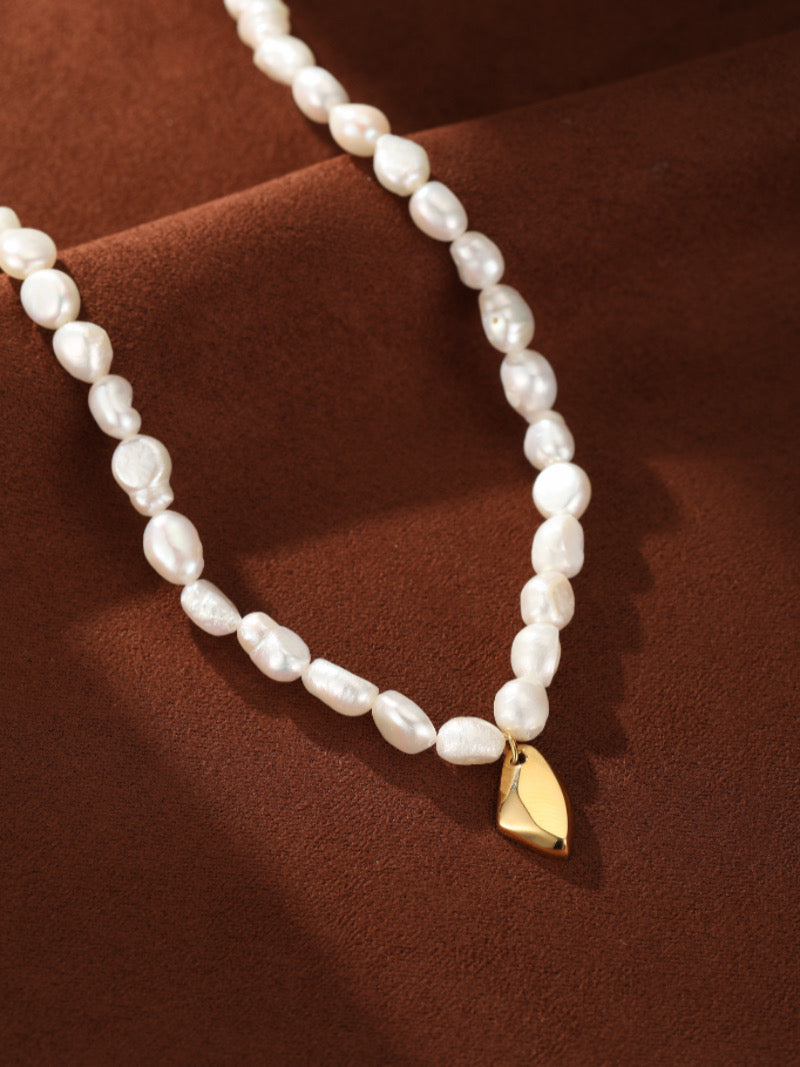Un collier ras-de-cou présentant un fil de perles blanches irrégulières d'eau douce et un pendentif goutte lisse et abstrait ton or sur une surface texturée marron.