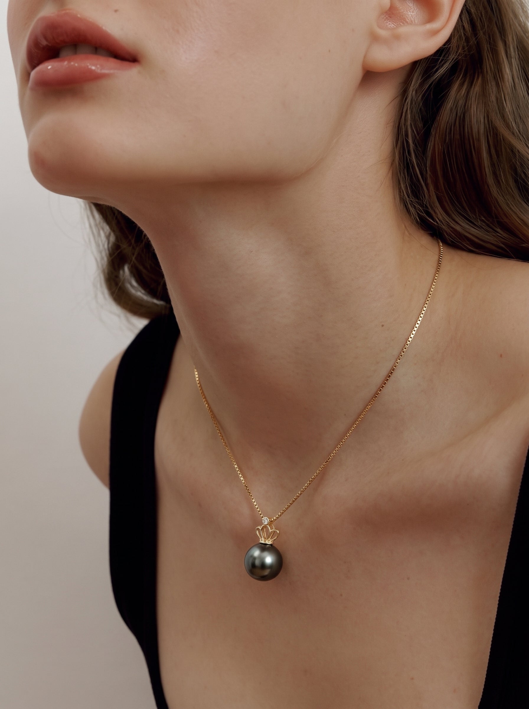 A close-up shot of a woman wearing the Majesty Tahitian Pearl Crown Pendant Necklace. The necklace features a single, lustrous 9mm round Tahitian black pearl suspended from a delicate gold chain, crowned with a subtle, sculptural motif and a tiny diamond accent. The pendant rests elegantly on the décolletage, visible beneath the neckline of her black top. @model
