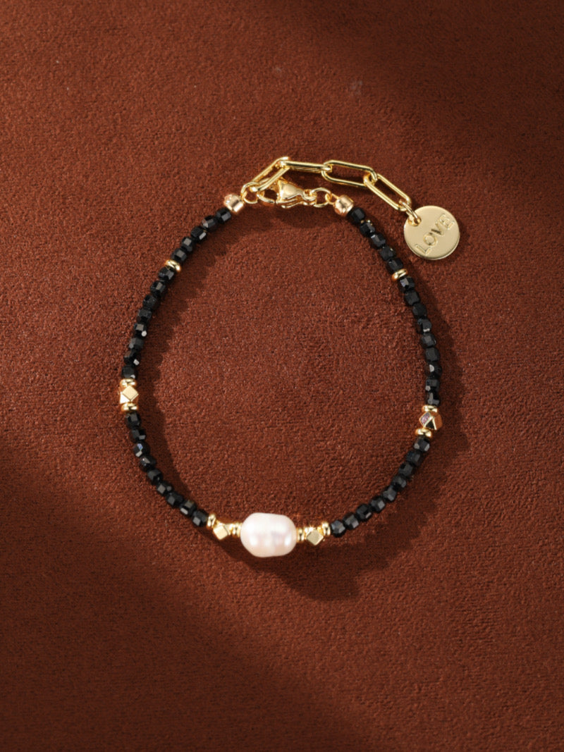 A black beaded bracelet with a single white baroque pearl and gold accents, featuring a small gold disc charm engraved with the word LOVE.
