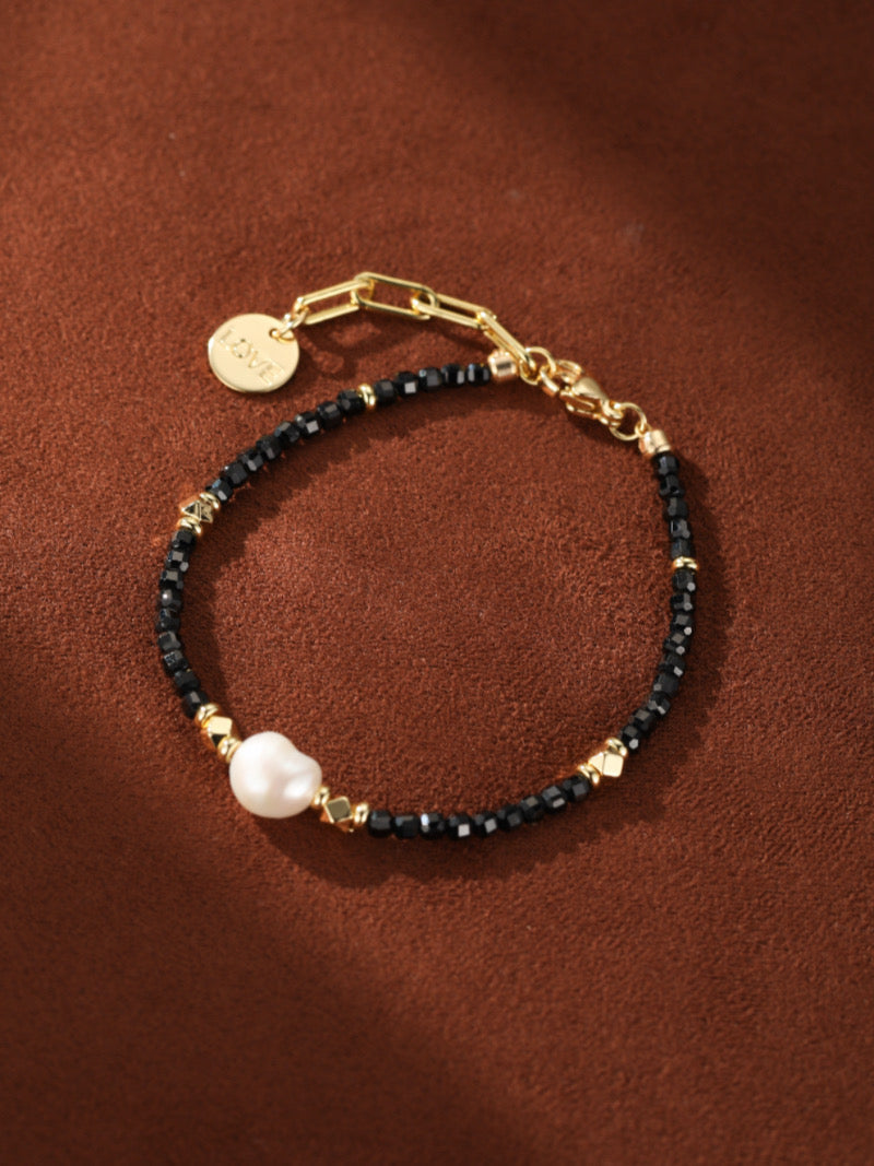 Matrix Freshwater Pearl Love Charm Chain Beaded Bracelet featuring a large white baroque pearl, faceted black beads, gold accents, and a "Love" charm on a brown background.