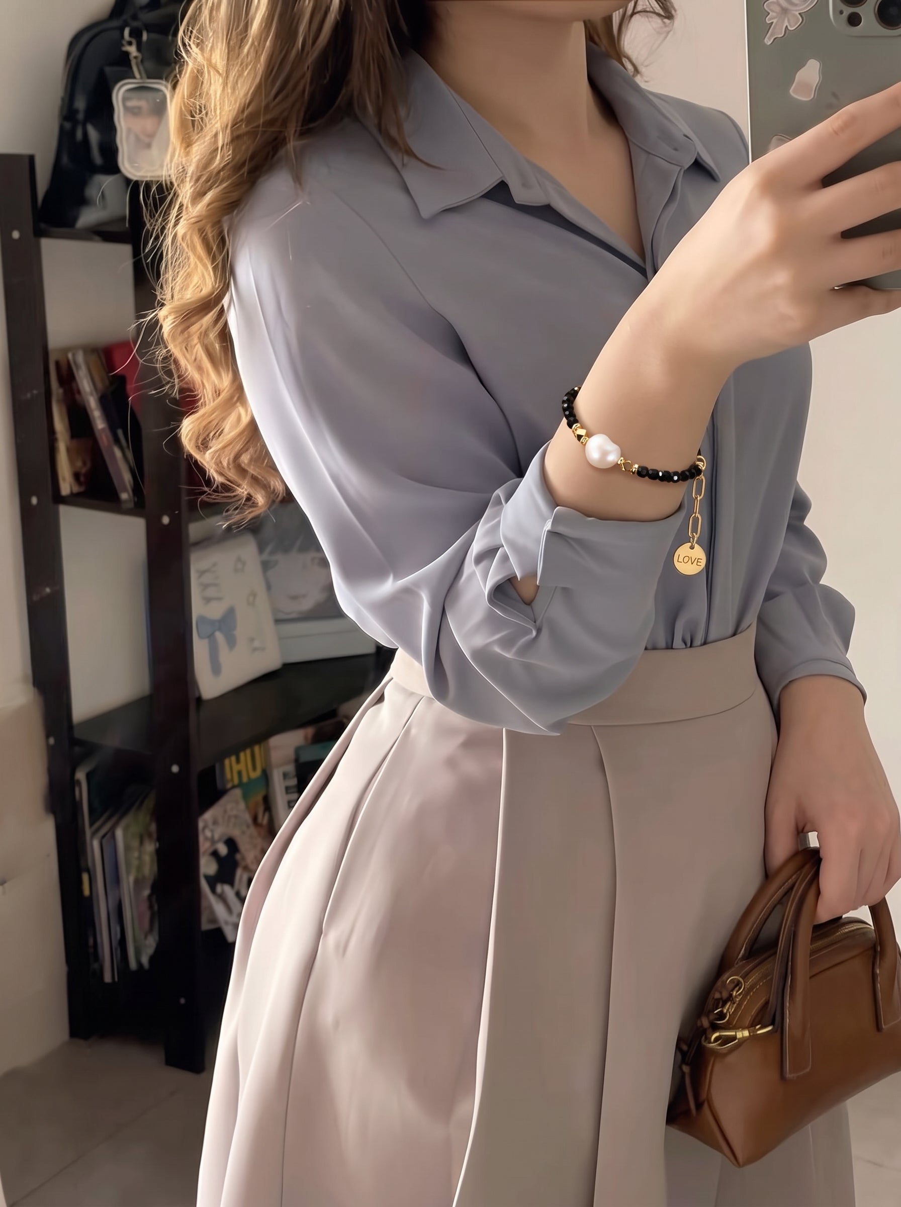 A woman wears the Matrix Freshwater Pearl Love Charm Chain Beaded Bracelet on her wrist, featuring a large white baroque pearl, black beads, gold accents, and a "love" charm. She is dressed in a light blue blouse and a beige skirt, holding a brown handbag. @lifestyle