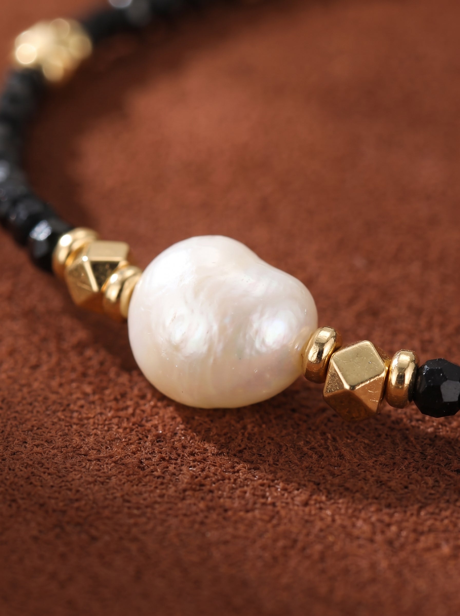 Close-up of a Matrix bracelet featuring a large white baroque freshwater pearl, faceted black beads, and gold-tone cube accents on a textured brown background. @pearl