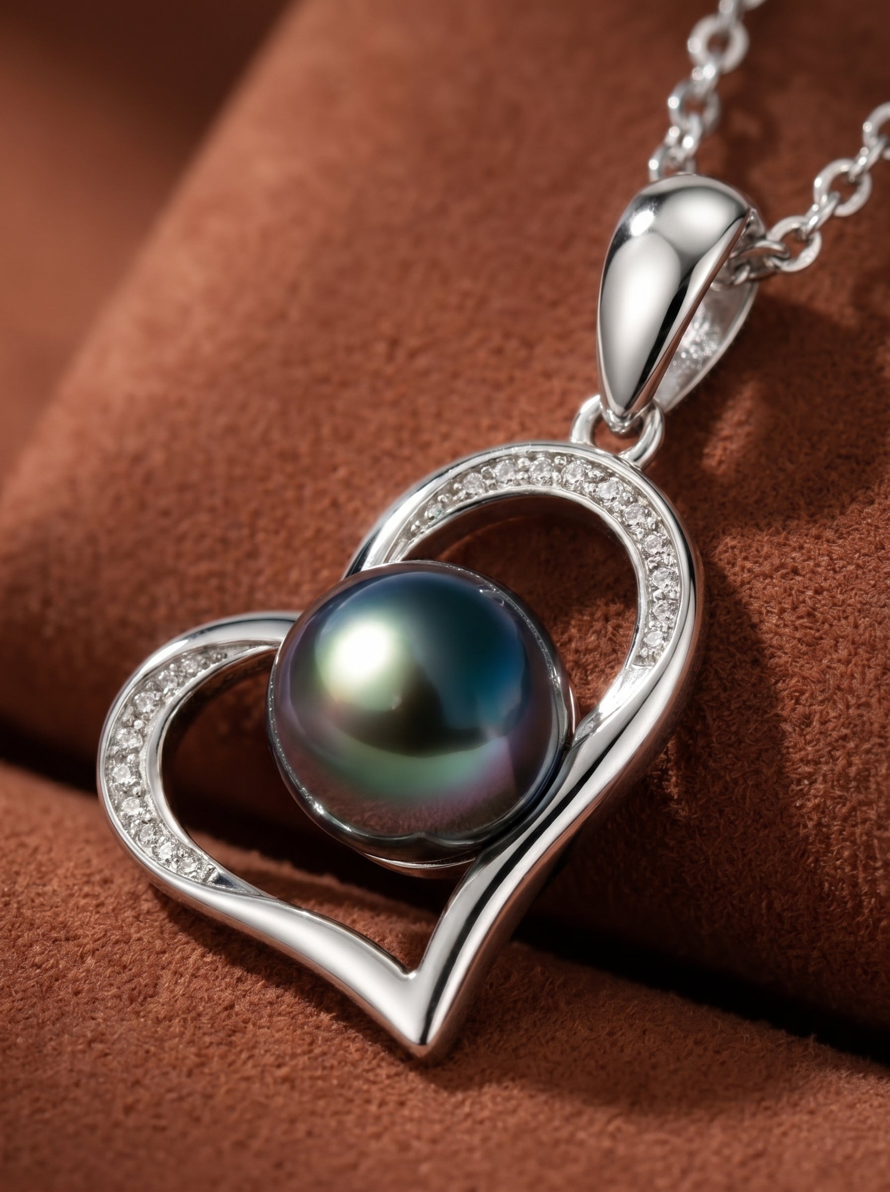 A silver heart pendant necklace with a single, iridescent Tahitian pearl at its center, accented with small diamonds. The pendant hangs from a delicate silver chain against a textured brown background.@pearl