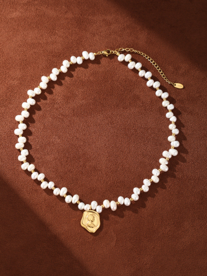 Collier pendentif perlé pièce Mythos en perle d'eau douce présentant de lustrées perles graines de 5 mm et un pendentif pièce doré sur fond marron.