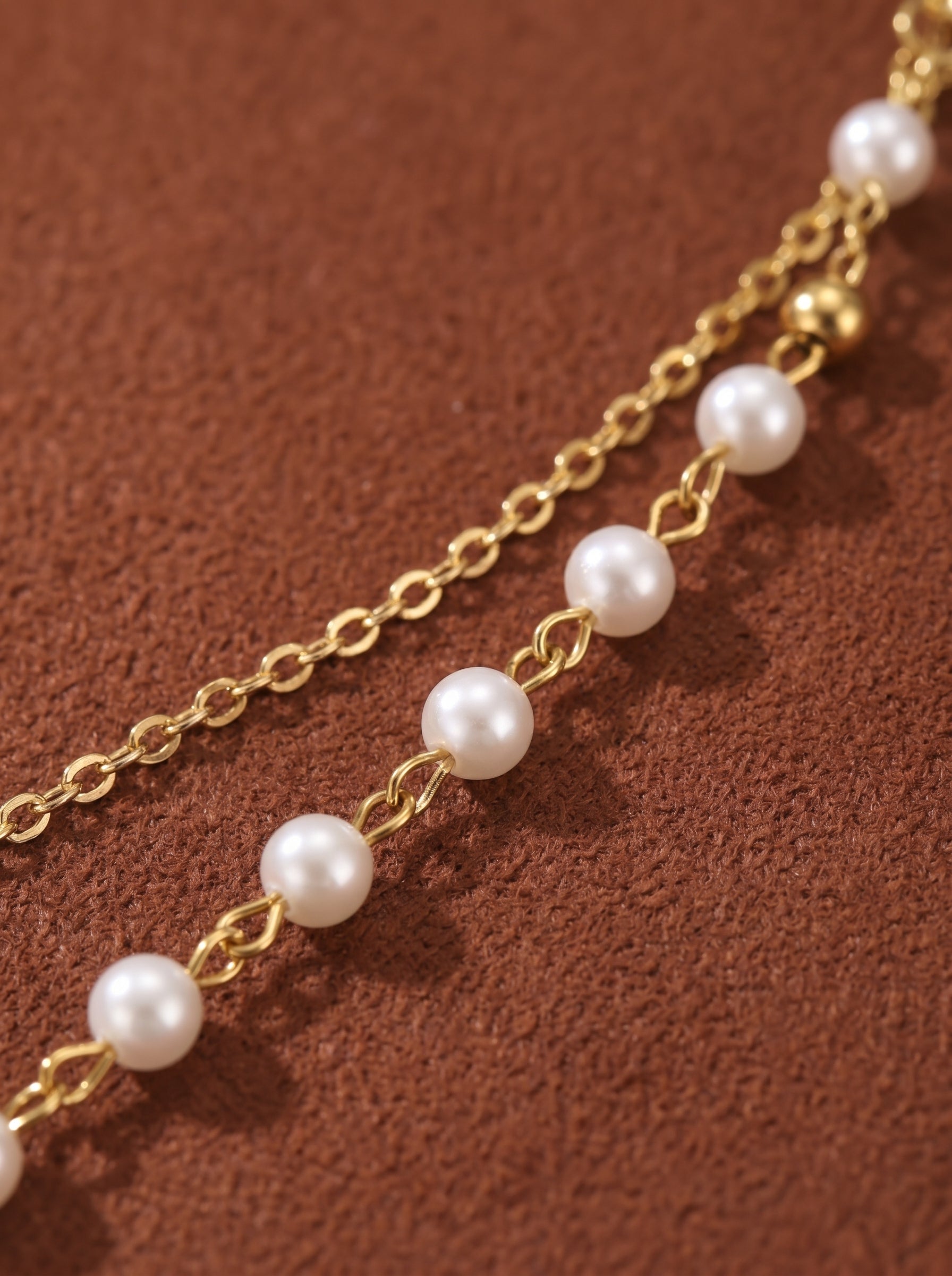 Close-up of a layered gold necklace with small white pearls and a gold bead on a textured brown background. @pearl