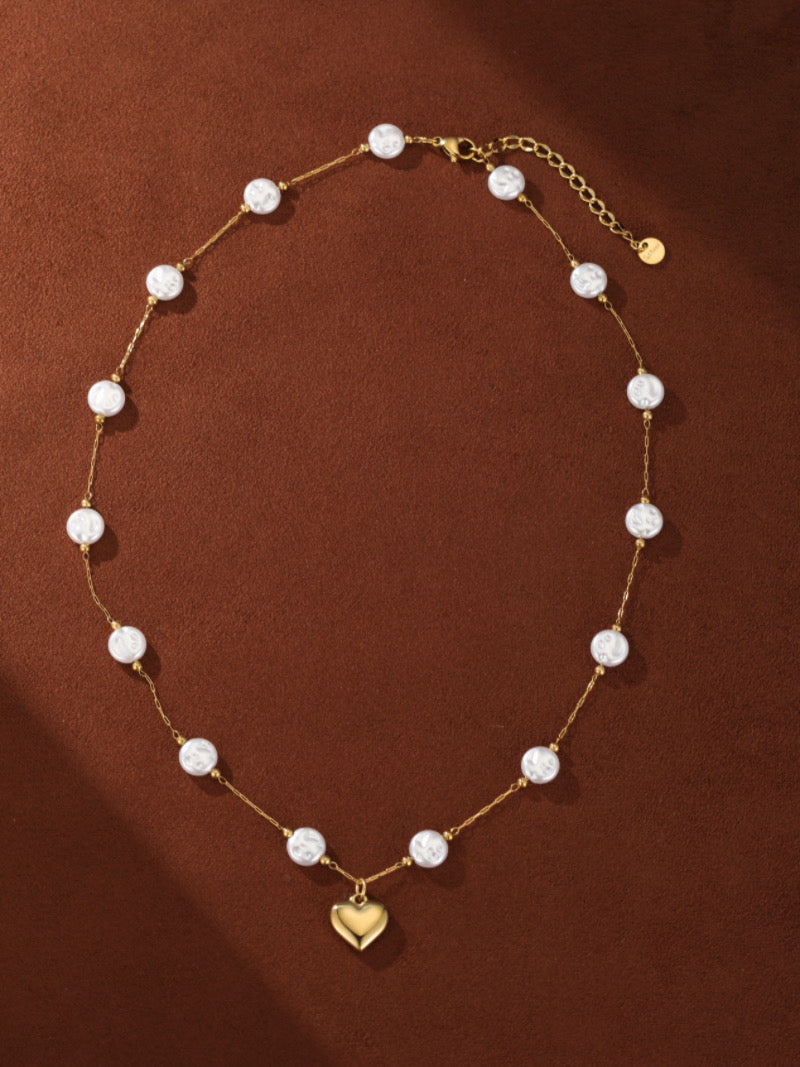 Gold-toned layered necklace with a delicate chain featuring round white pearls and a small, shiny gold heart pendant at the bottom, on a brown background.