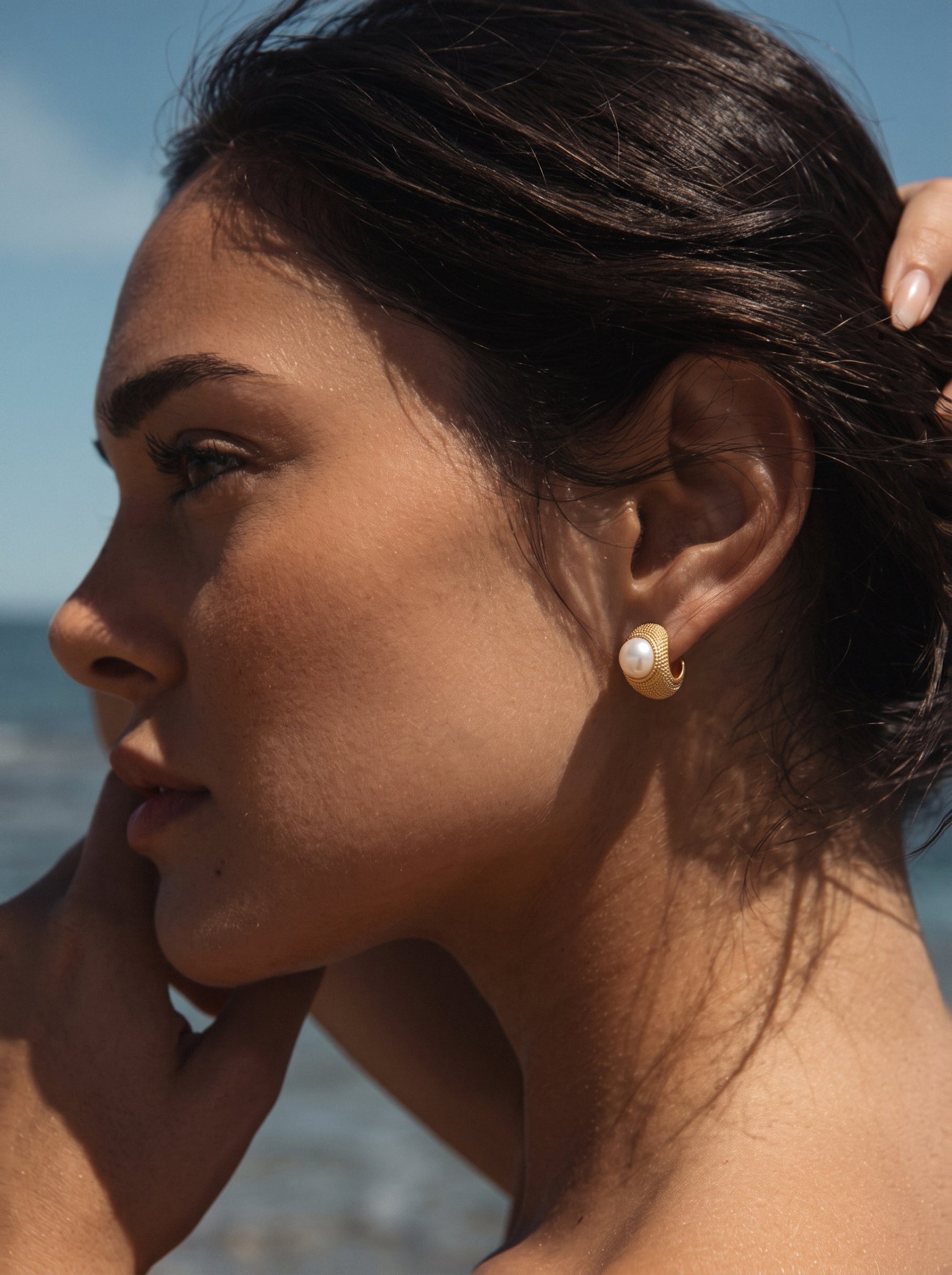 Close-up of a woman wearing Orbit freshwater pearl textured stud earrings with a textured gold-tone setting against a blurred blue ocean background. @model #lifestyle