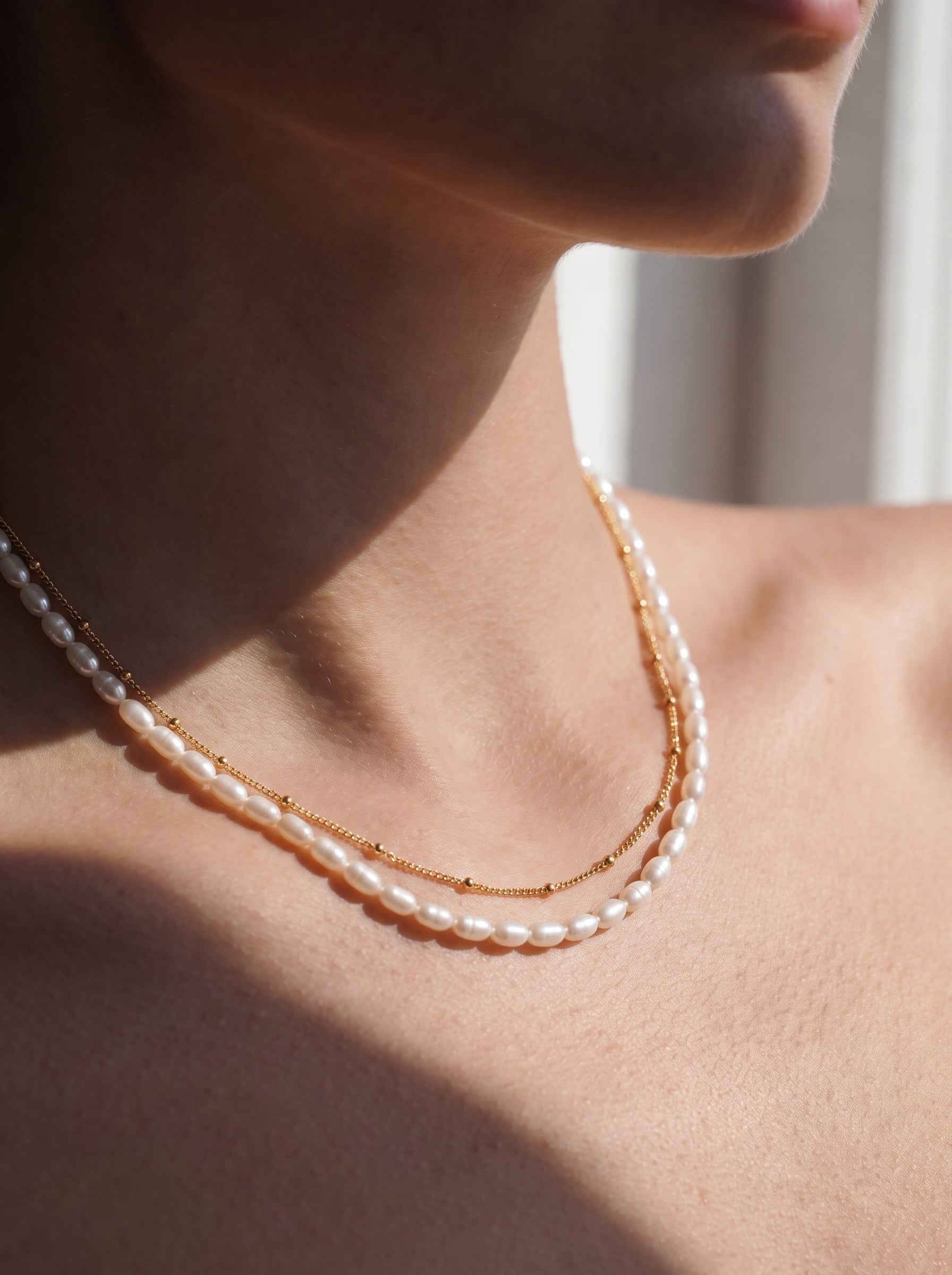 Un collier superposé avec un fil de perles blanches en forme graine d'eau douce et une délicate chaîne dorée avec de petites perles, porté par une mannequin. @model