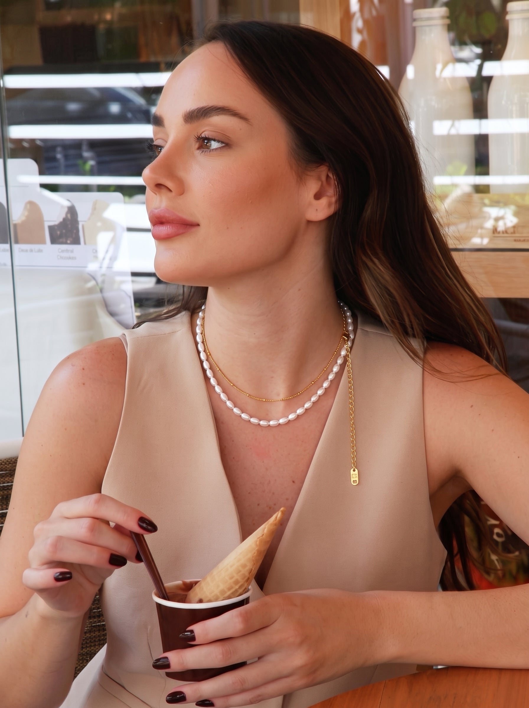 Une femme en haut beige portant un collier superposé avec un fil de perles graines blanches d'eau douce et une délicate chaîne dorée, tenant une coupe de glace. @lifestyle