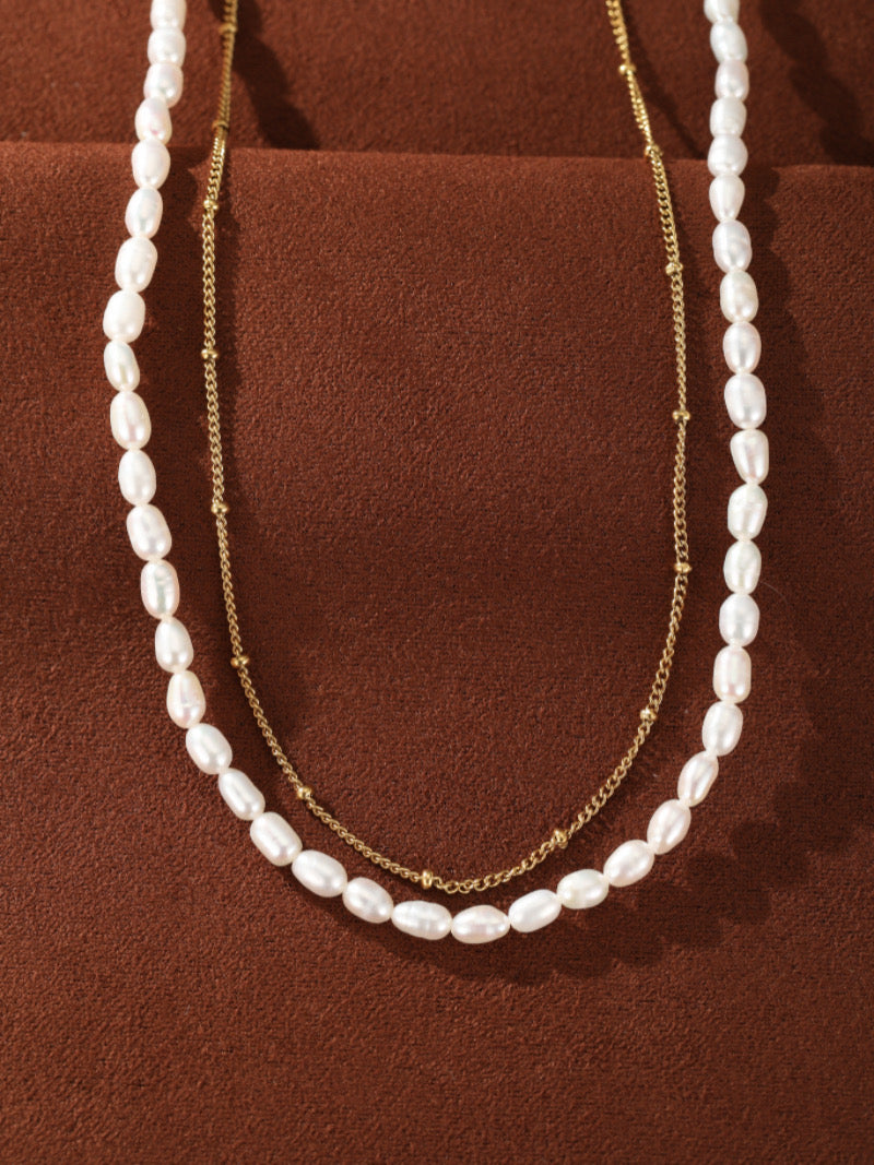 Un collier superposé présentant un fil de perles blanches en forme graine d'eau douce et une délicate chaîne ton or avec de petits accents de perle.