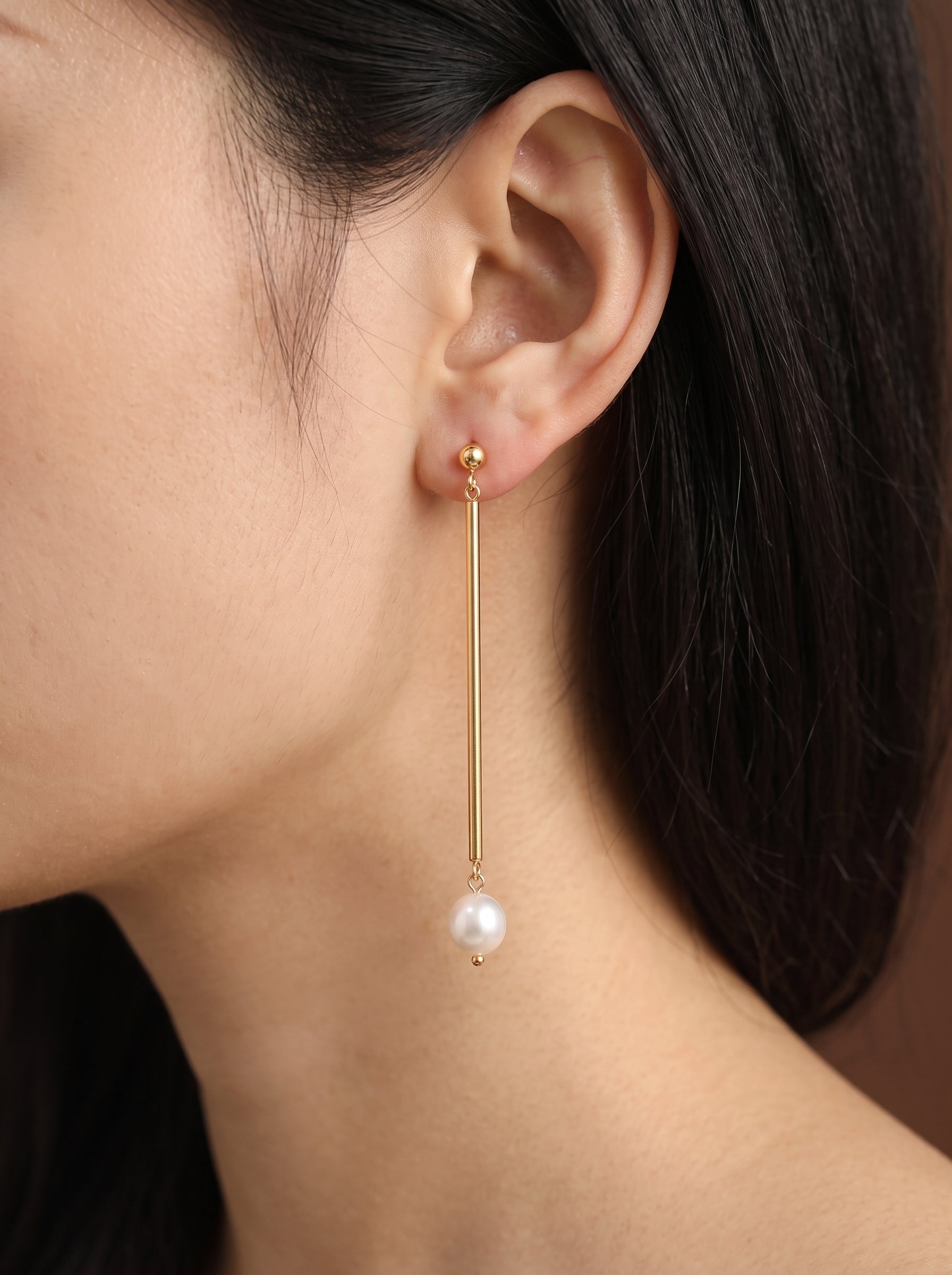 Gros plan d'une mannequin portant des boucles d'oreilles goutte Pillar ton or avec une seule perle blanche d'eau douce pendant d'une fine barre. La boucle d'oreille est montrée à son oreille, mettant en valeur son design élégant et minimaliste. @model