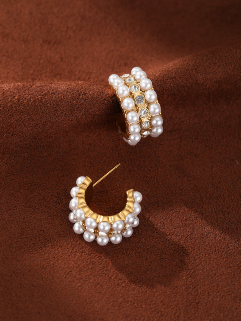 A pair of gold hoop earrings adorned with multiple rows of small white freshwater pearls and clear crystals, set against a textured brown background.