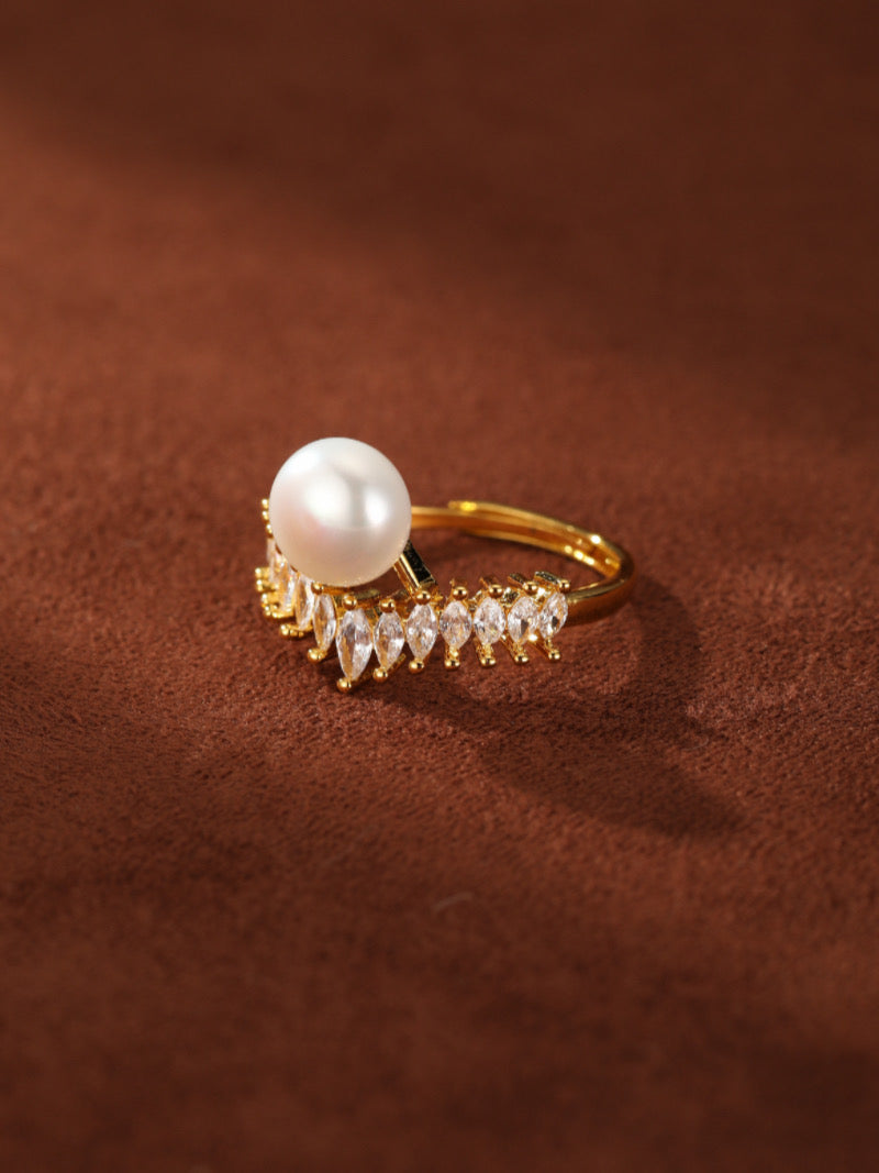 A gold cuff ring featuring a large white freshwater pearl centered above a row of sparkling marquise-cut cubic zirconia stones, set against a warm brown background.