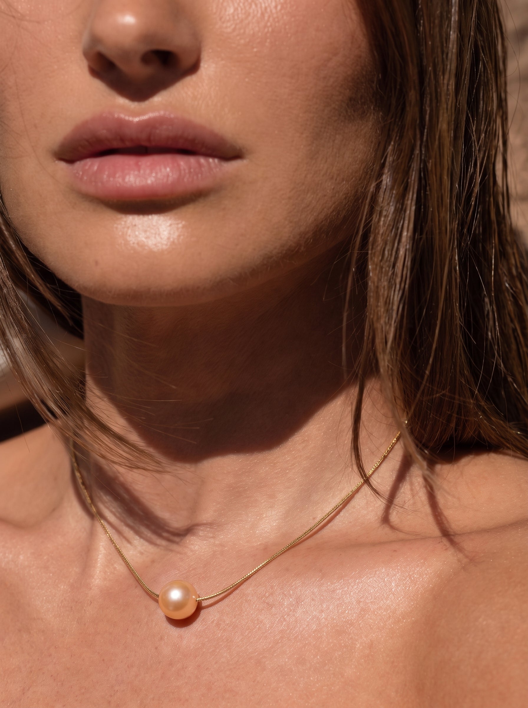 Gros plan d'une femme portant le collier pendentif goutte unique Poise en perle d'eau douce avec une seule perle couleur pêche sur une délicate chaîne dorée. @model