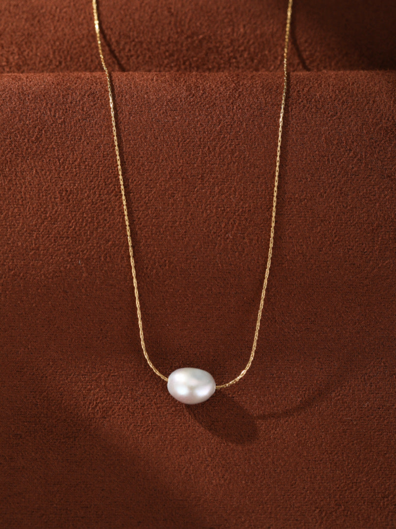 Collier Poise avec une seule perle blanche d'eau douce suspendue à une délicate chaîne dorée contre un fond texturé marron.