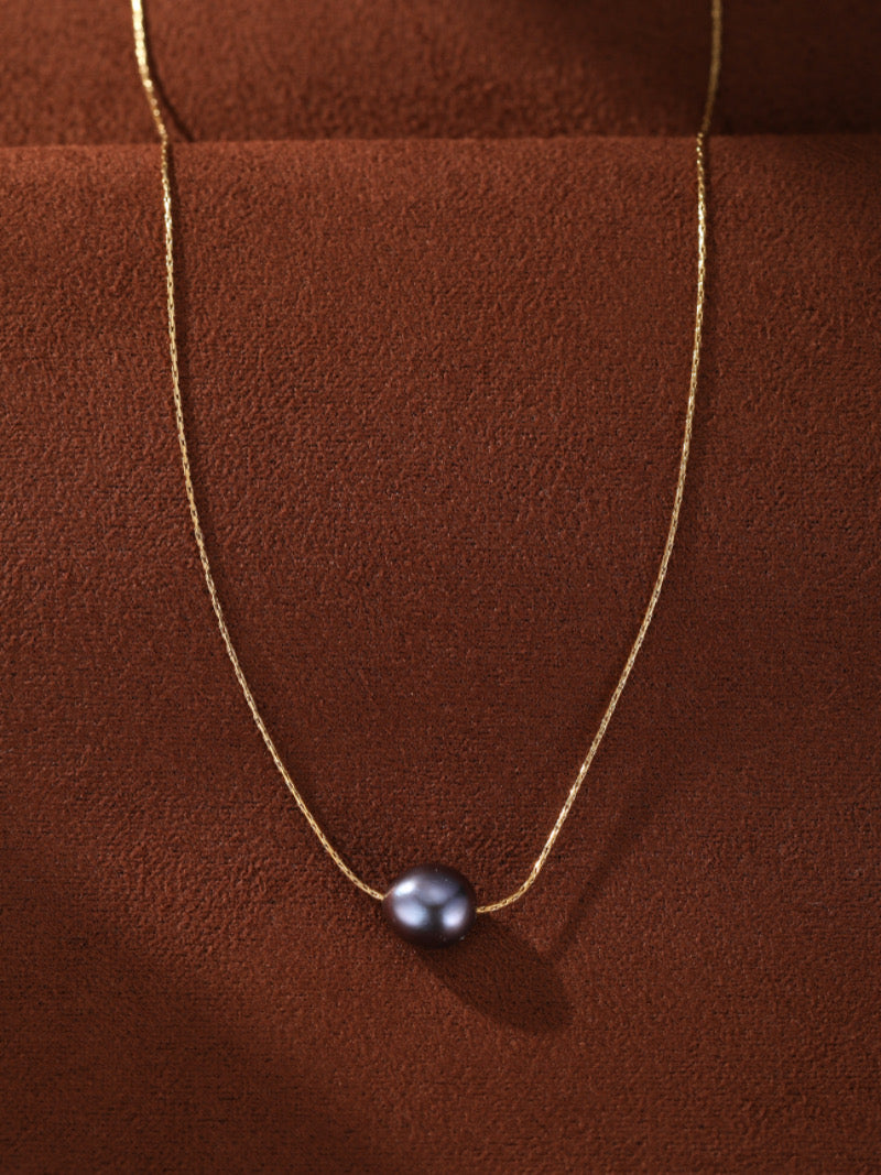 Collier Poise présentant une seule perle noire d'eau douce suspendue à une délicate chaîne dorée contre un fond texturé marron riche.