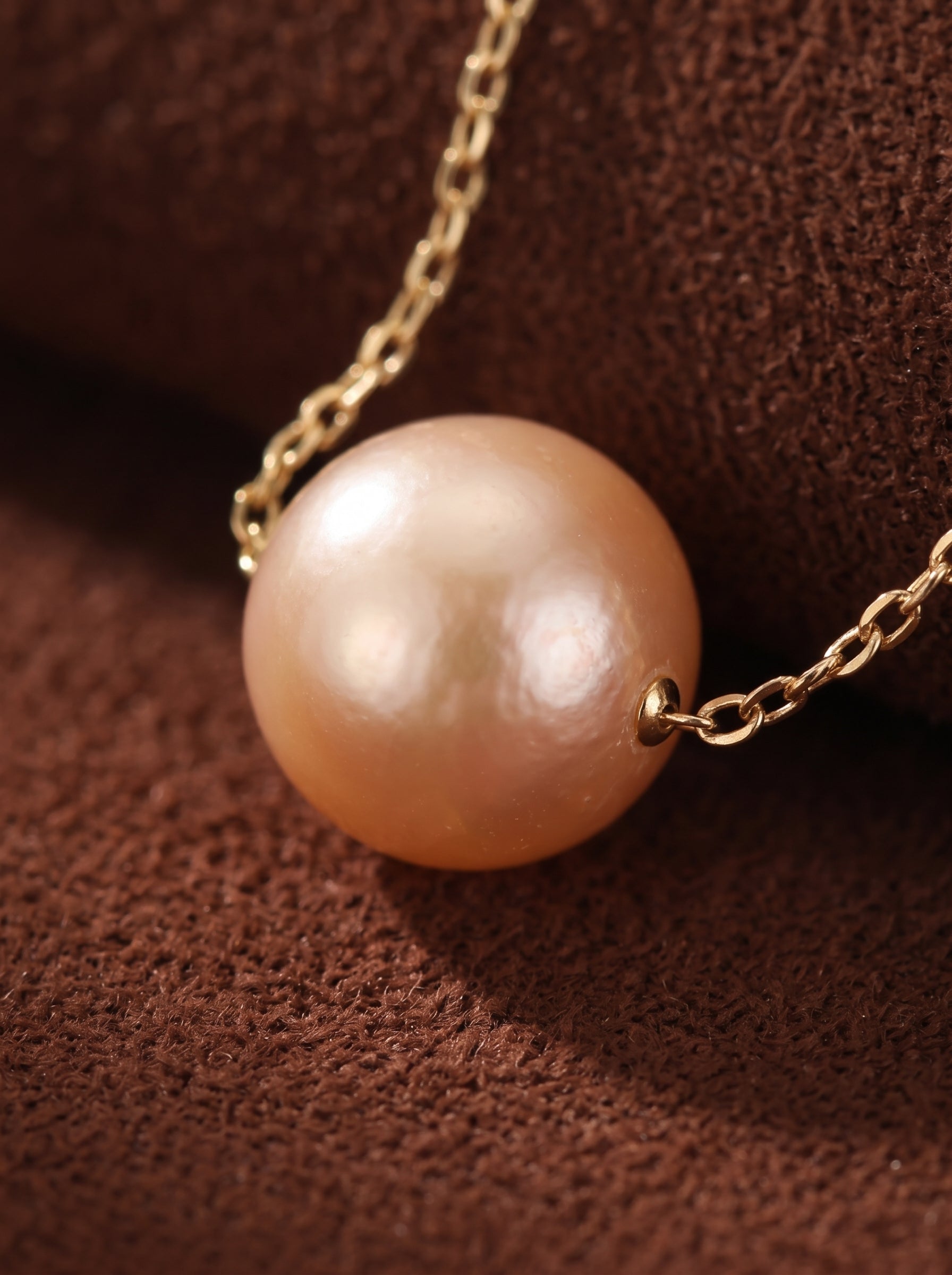 Collier Poise présentant une seule perle pêche d'eau douce suspendue à une délicate chaîne dorée contre un fond texturé marron. @pearl