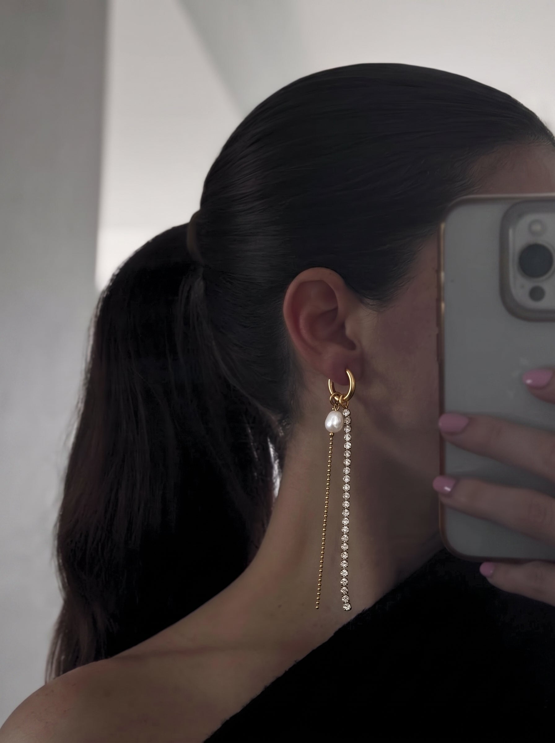 Close-up of a woman's ear wearing a gold hoop earring with a dangling freshwater pearl and a long chain of crystals. @model