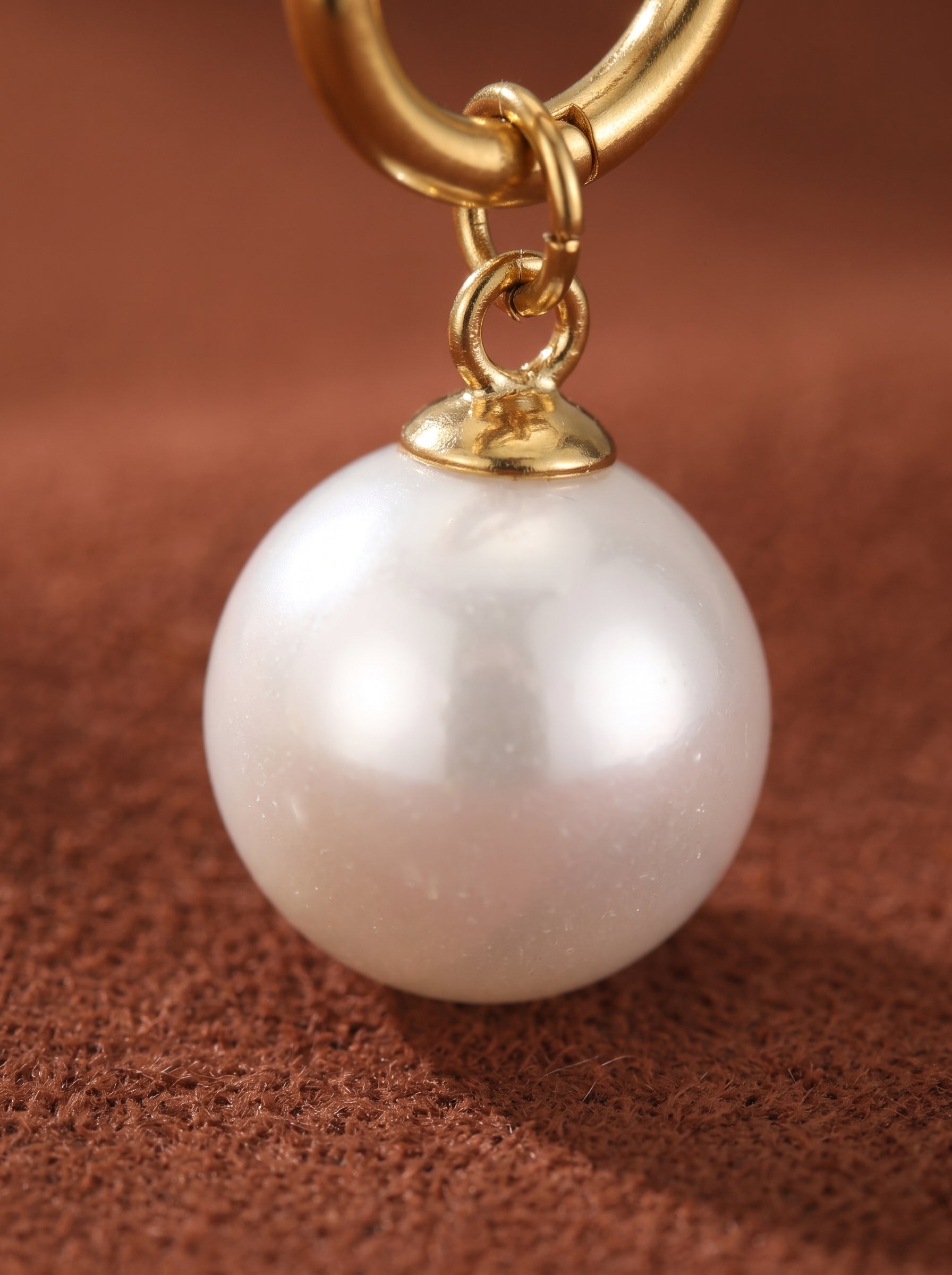 Gros plan d'une créole dorée avec une seule et lumineuse perle pendante blanche d'eau douce de 8 mm suspendue. La perle est ronde et lisse, révélant un éclat naturel contre un fond texturé marron. @perle