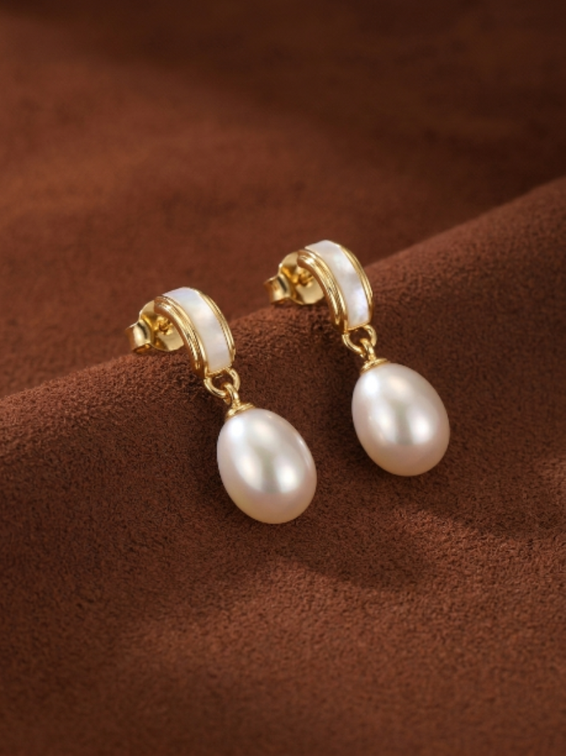 A pair of Quietude Freshwater Pearl Drop Earrings featuring gold-toned curved elements with white inlay and luminous 6mm seed-shaped white pearls dangling below, set against a brown textured background.