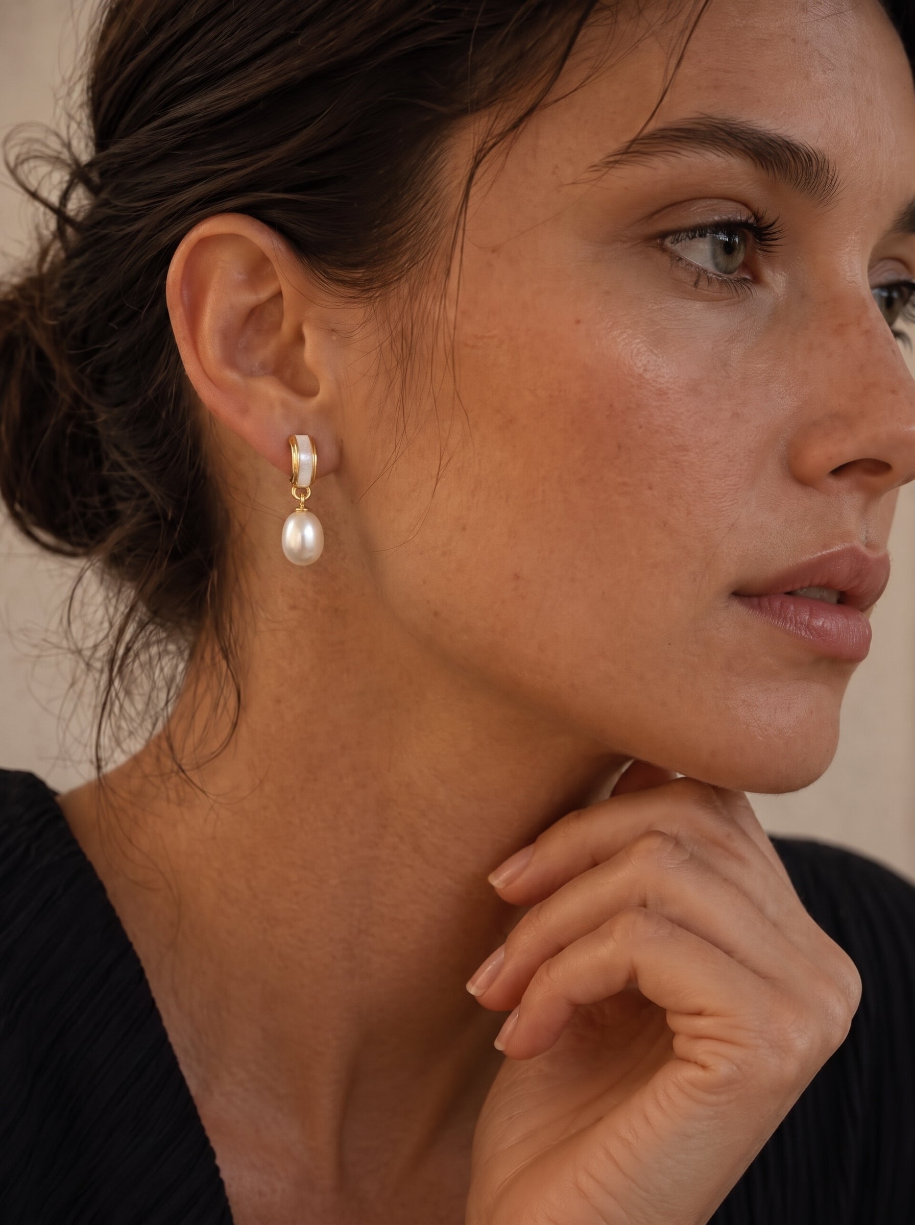 A woman wearing Quietude freshwater pearl drop earrings, featuring a polished gold-toned curved element with a white inlay and a single luminous seed-shaped white pearl drop. The earrings are positioned on her ear, showcasing their elegant design. @lifestyle