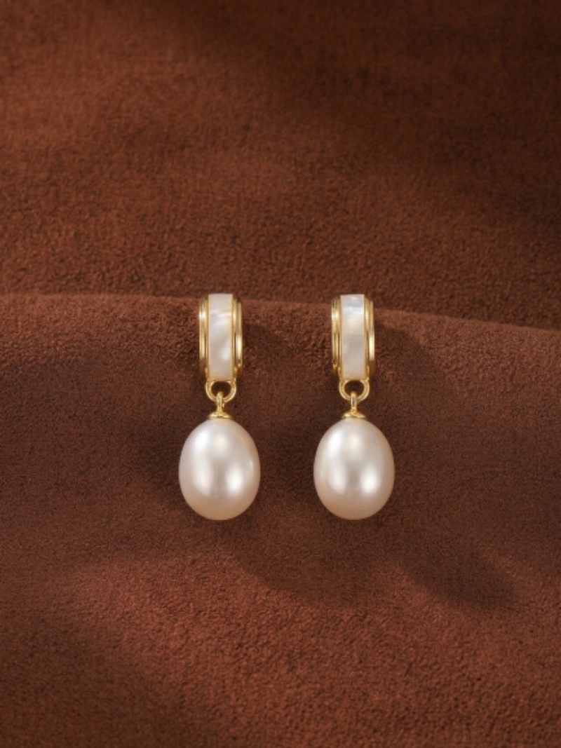 Pair of gold-toned Quietude freshwater pearl drop earrings with a curved element and a single luminous white seed pearl hanging below, set against a textured brown background.