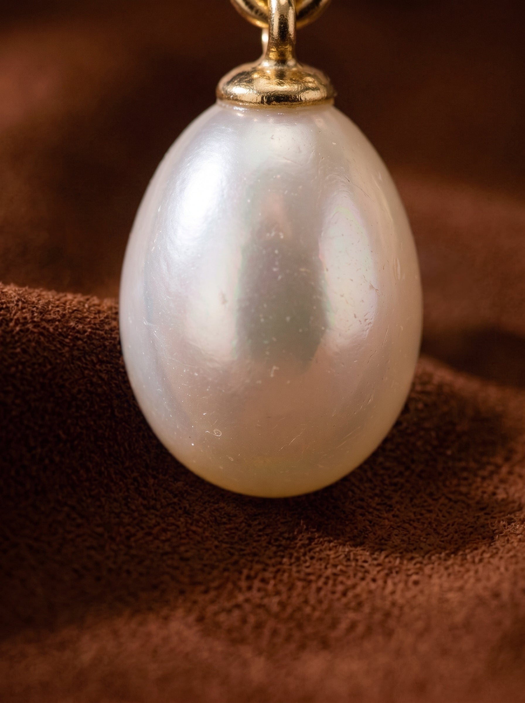 A close-up shot of a single, luminous 6mm seed-shaped Freshwater Pearl earring, showcasing its smooth, iridescent surface and the delicate gold-toned cap it hangs from. The pearl rests against a textured brown fabric background. @pearl