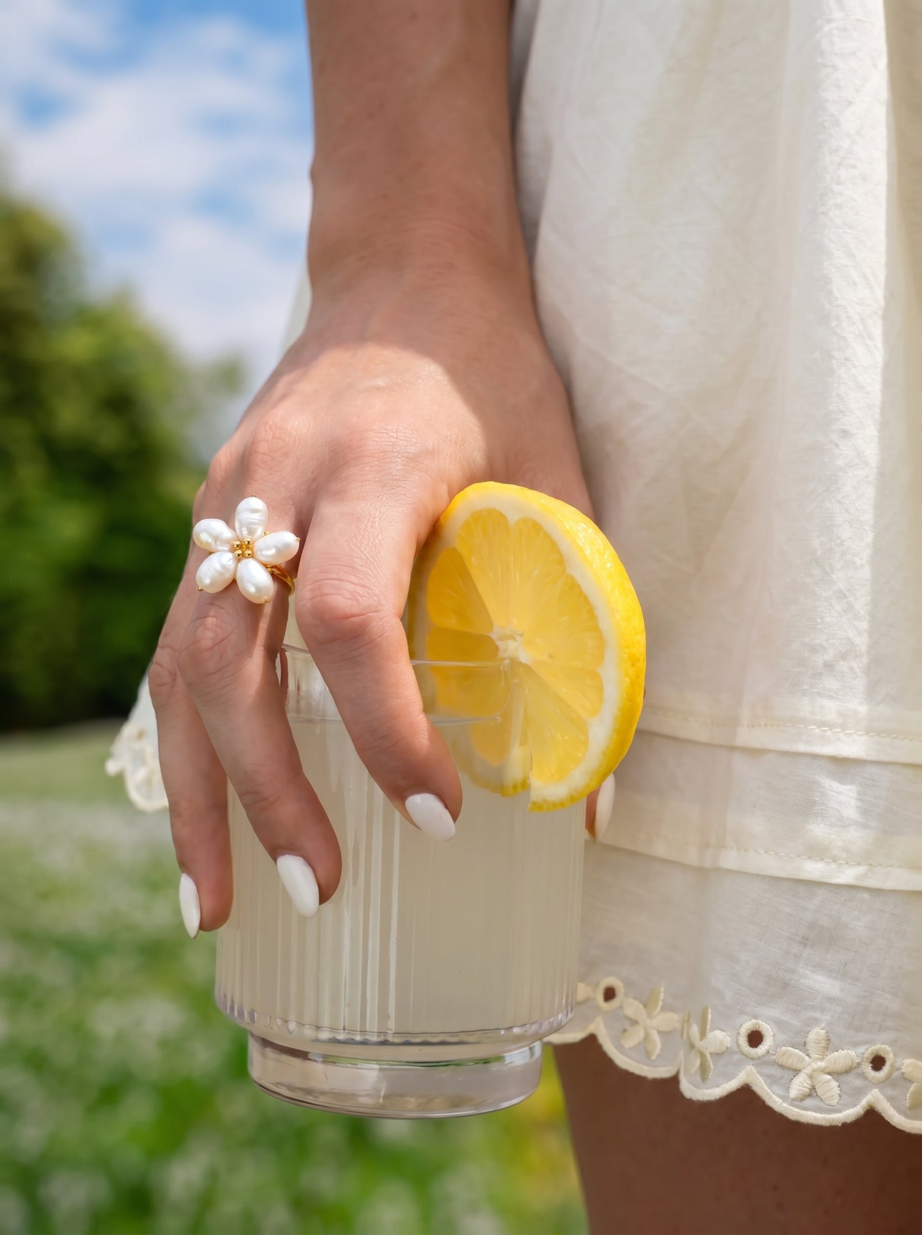 A hand wearing a Radiance freshwater pearl flower cuff ring holds a glass of lemonade with a lemon slice, set against a backdrop of a sunny green meadow with a blue sky. @lifestyle #lifestyle