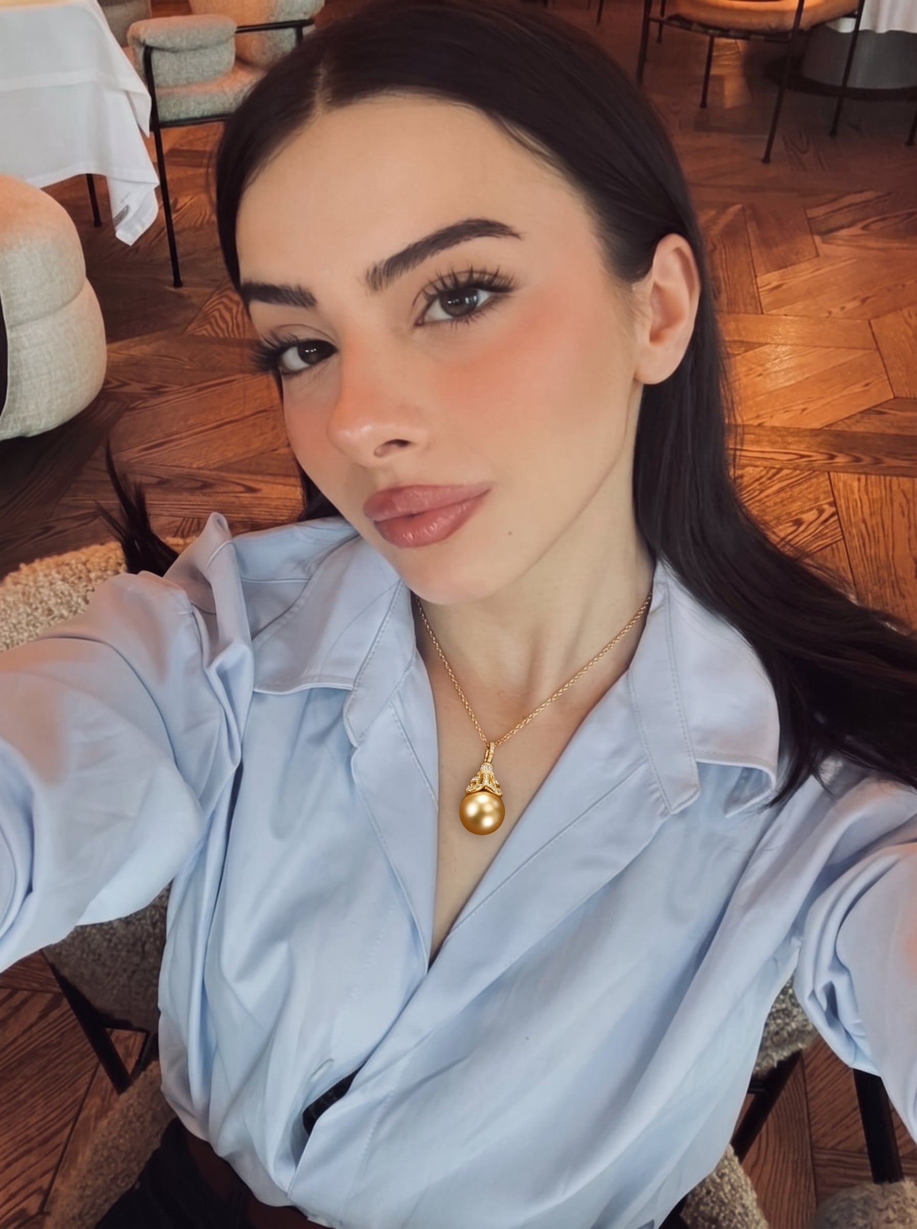 A woman wearing a light blue collared shirt and a gold pendant necklace featuring a large, lustrous South Sea pearl with a diamond-encrusted cap. The necklace is the focal point of the image, worn in a professional or casual setting. @lifestyle