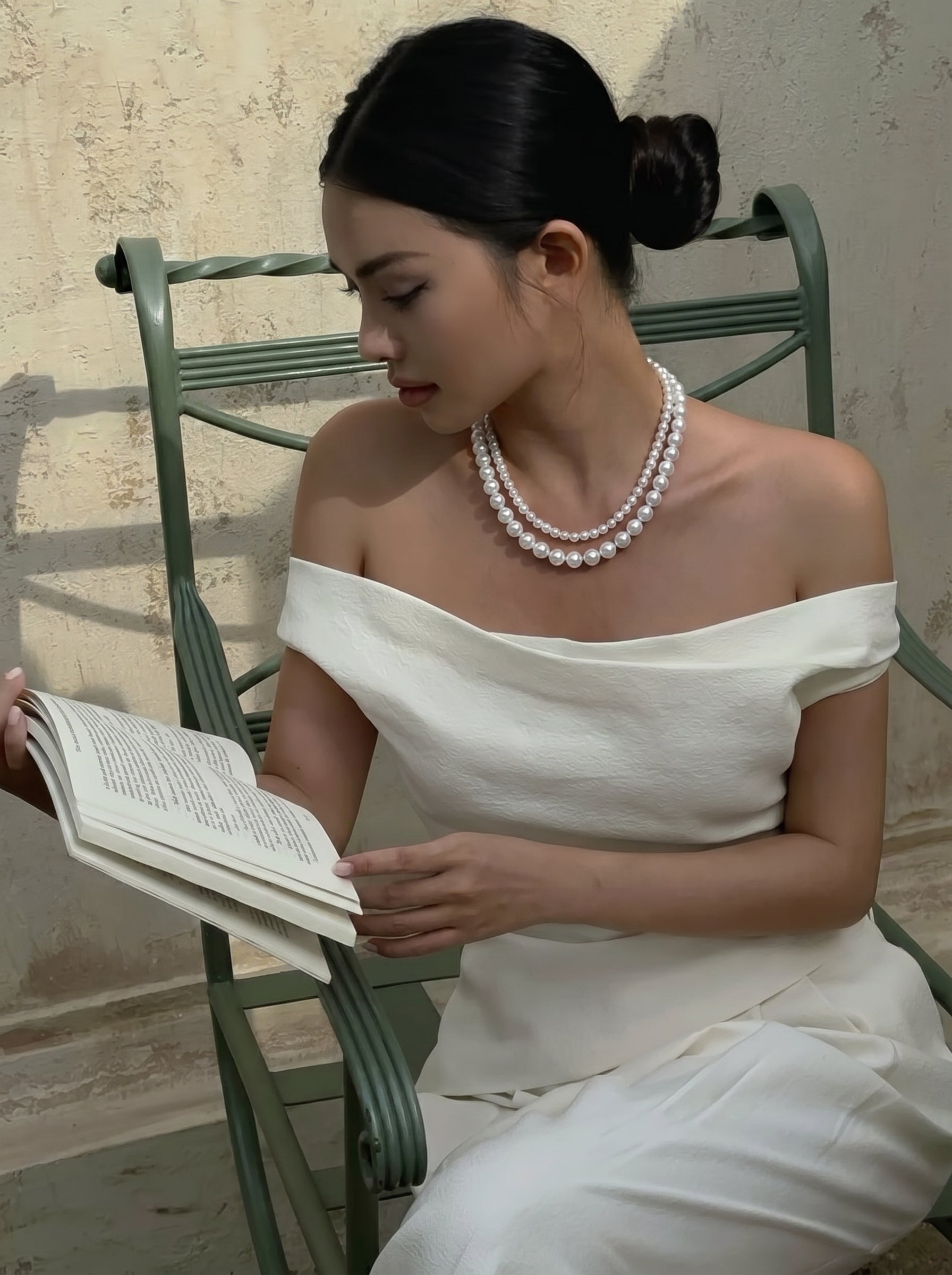 A woman wearing a white off-the-shoulder top and the Rhapsody Freshwater Pearl Layered Necklace, sitting on a green chair and reading a book. @lifestyle #lifestyle