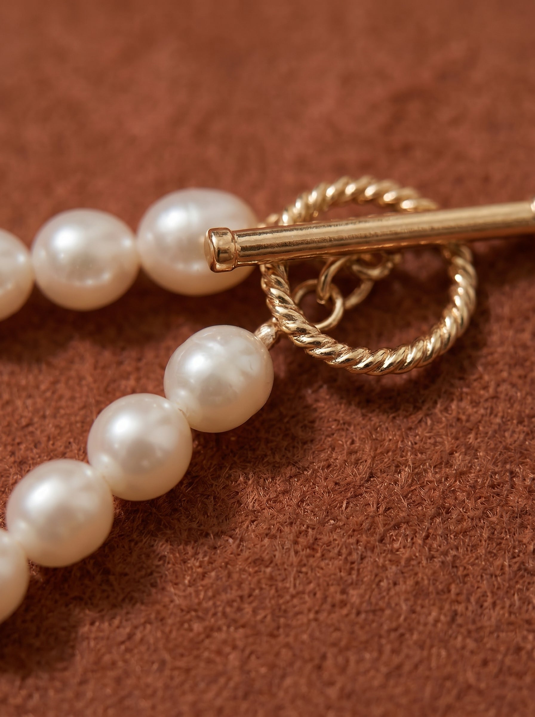 Close-up of a long freshwater pearl necklace with a gold-tone twisted toggle clasp on a textured brown background. @pearl
