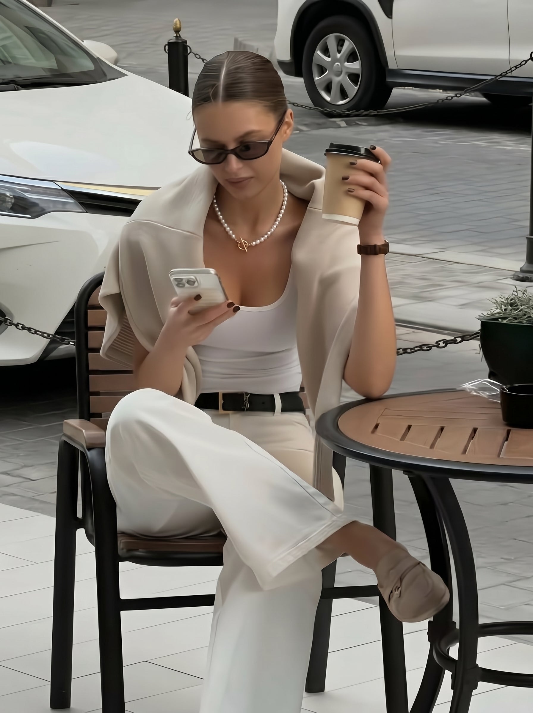 A woman wearing a white tank top and cream cardigan over her shoulders, with white wide-leg pants, sits at an outdoor cafe table holding a coffee cup and looking at her phone. She is wearing the Rhythm Freshwater Pearl Long Necklace with a gold clasp and pearl earrings. @lifestyle #lifestyle