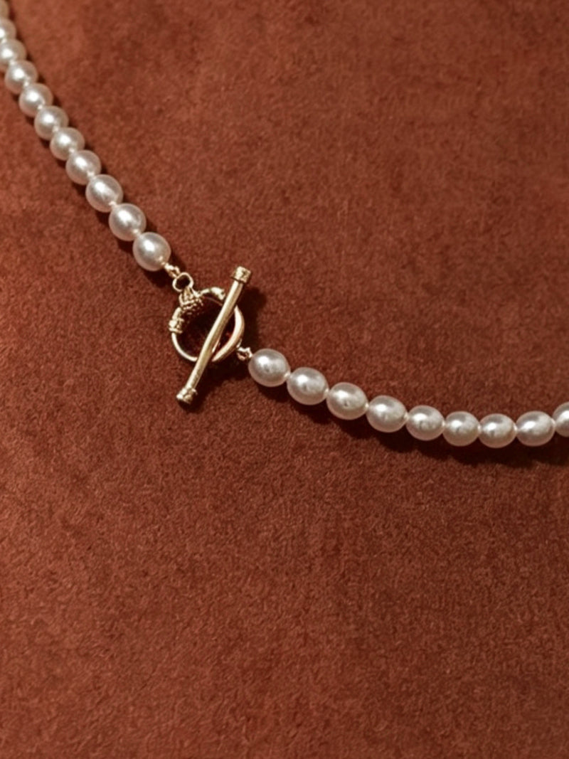 Close up of a long, delicate white freshwater pearl necklace with a gold toggle clasp against a textured brown background.