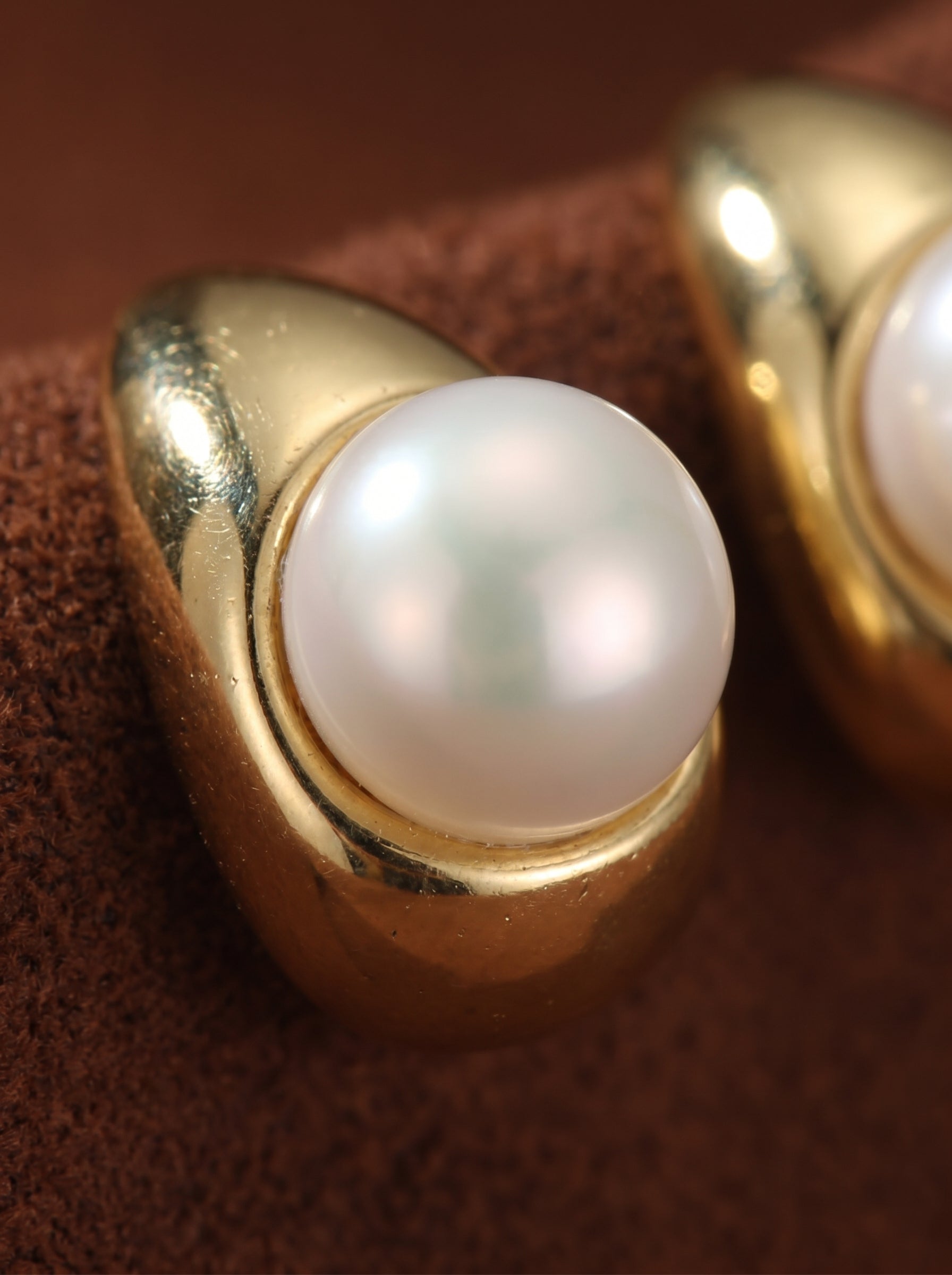 Close-up of a gold-toned Sanctuary moon stud earring featuring a luminous white button freshwater pearl. The pearl is set in a sleek, modern gold casing against a textured brown background. @pearl