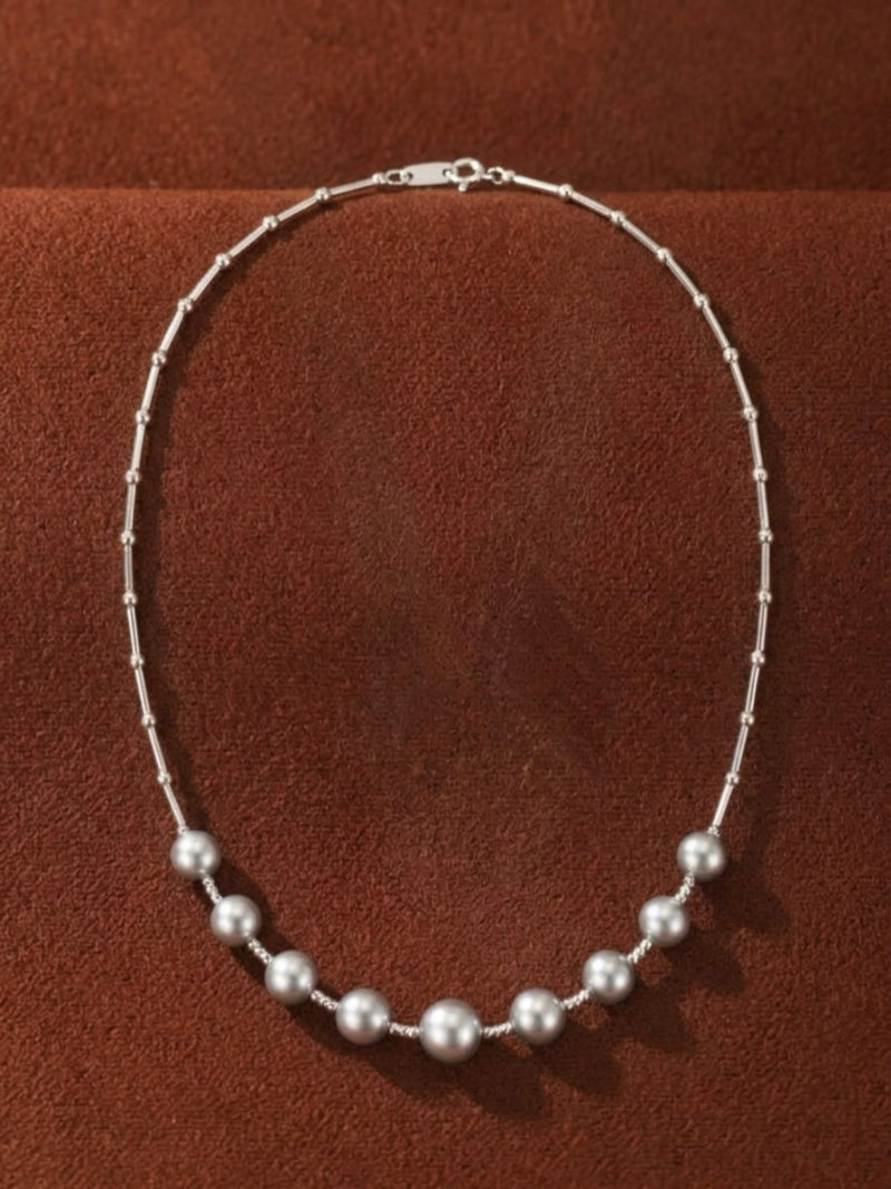 A delicate silver necklace featuring a scattering of luminous Akoya pearls on a beaded chain against a textured brown background.