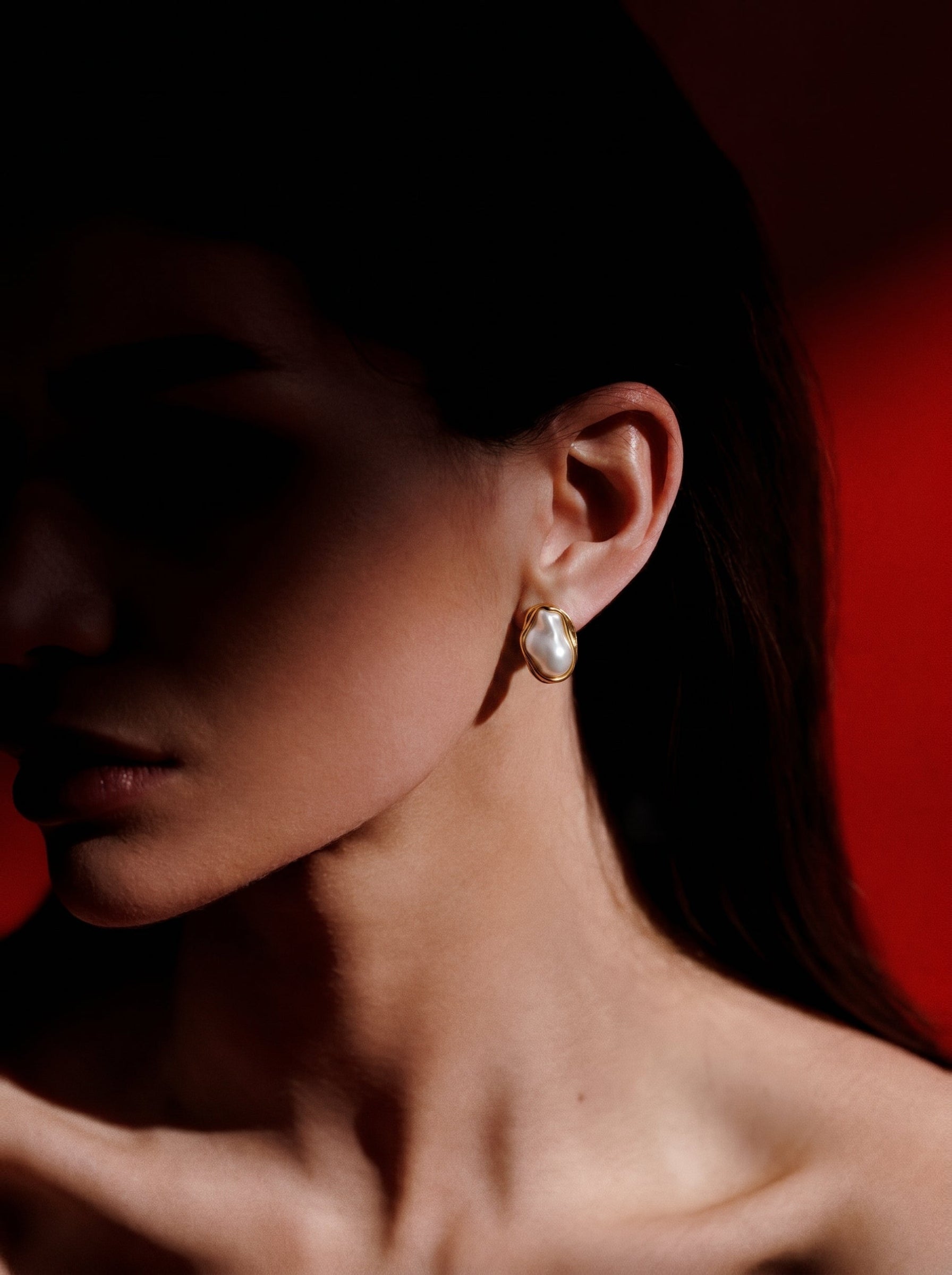 A close-up shot of a woman's ear showcasing a Sculpted freshwater pearl framed stud earring. The earring features a large, irregular white baroque pearl with a lustrous sheen, set within a fluid, golden-toned frame. The background is a vibrant red, with dramatic lighting creating shadows on the woman's face and neck. @model