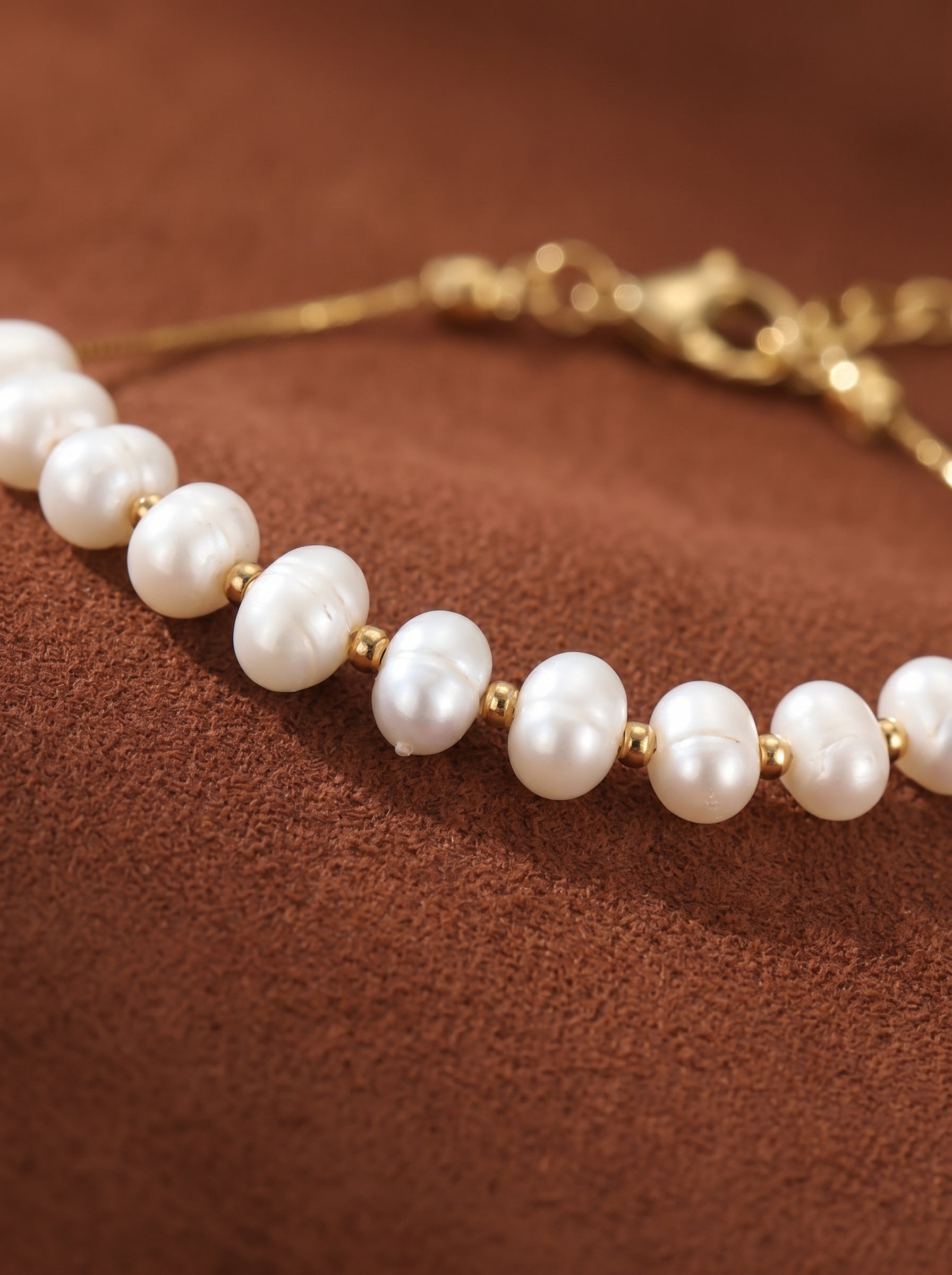 A delicate gold chain bracelet features a row of luminous 7mm freshwater pearls interspersed with small gold beads, set against a textured brown background. @pearl