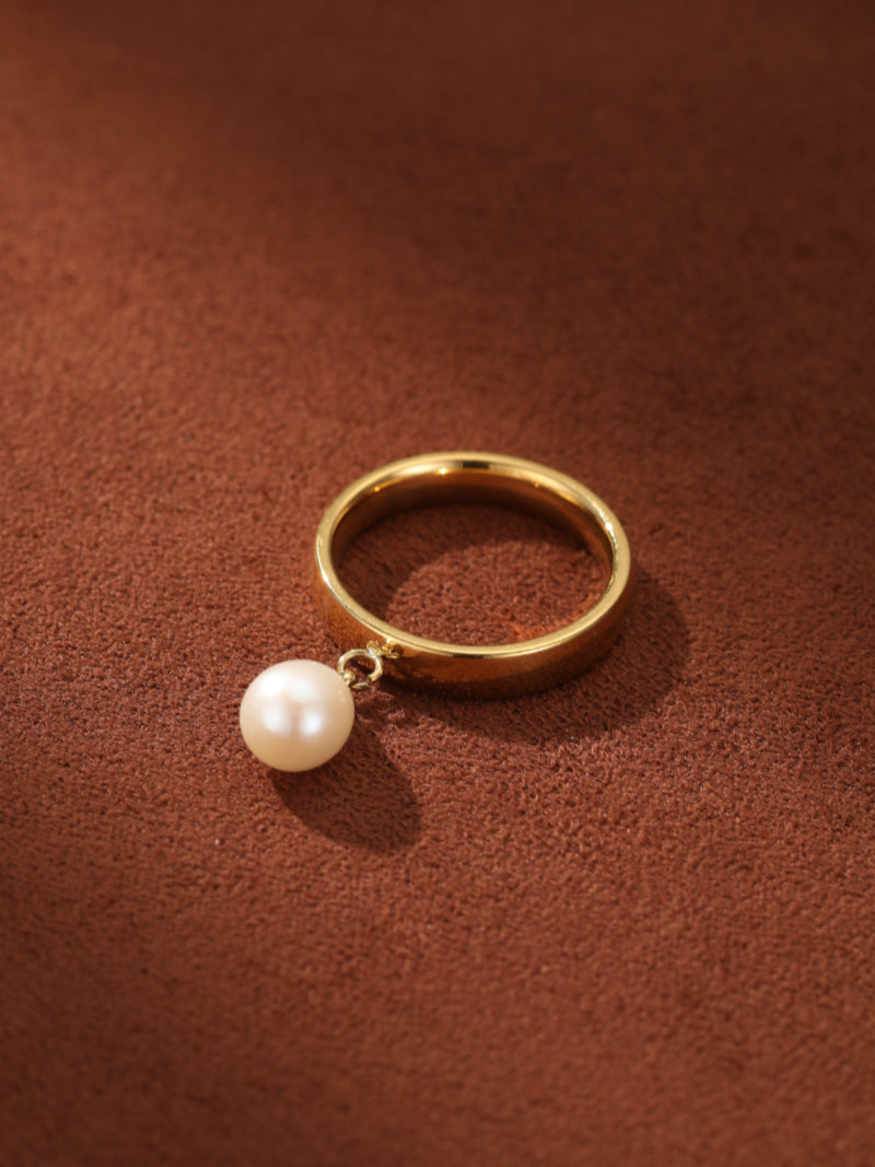 Gold ring with a single white freshwater pearl suspended from a delicate bead, set against a textured brown surface.@pearl