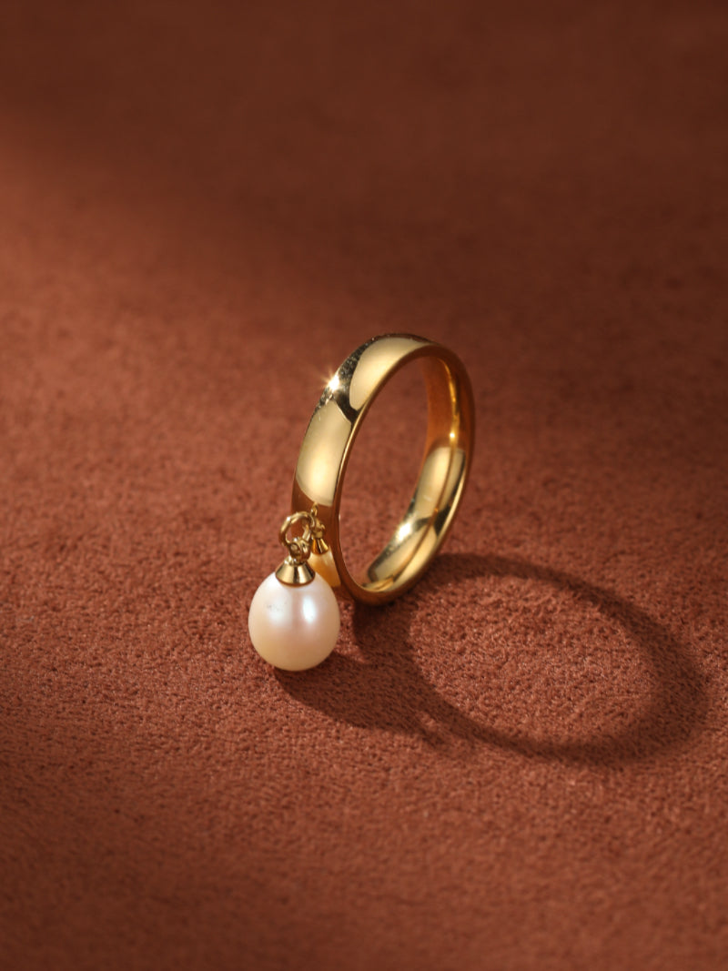 A minimalist gold ring with a single, lustrous white freshwater pearl dangling from a delicate chain attached to the band. @pearl