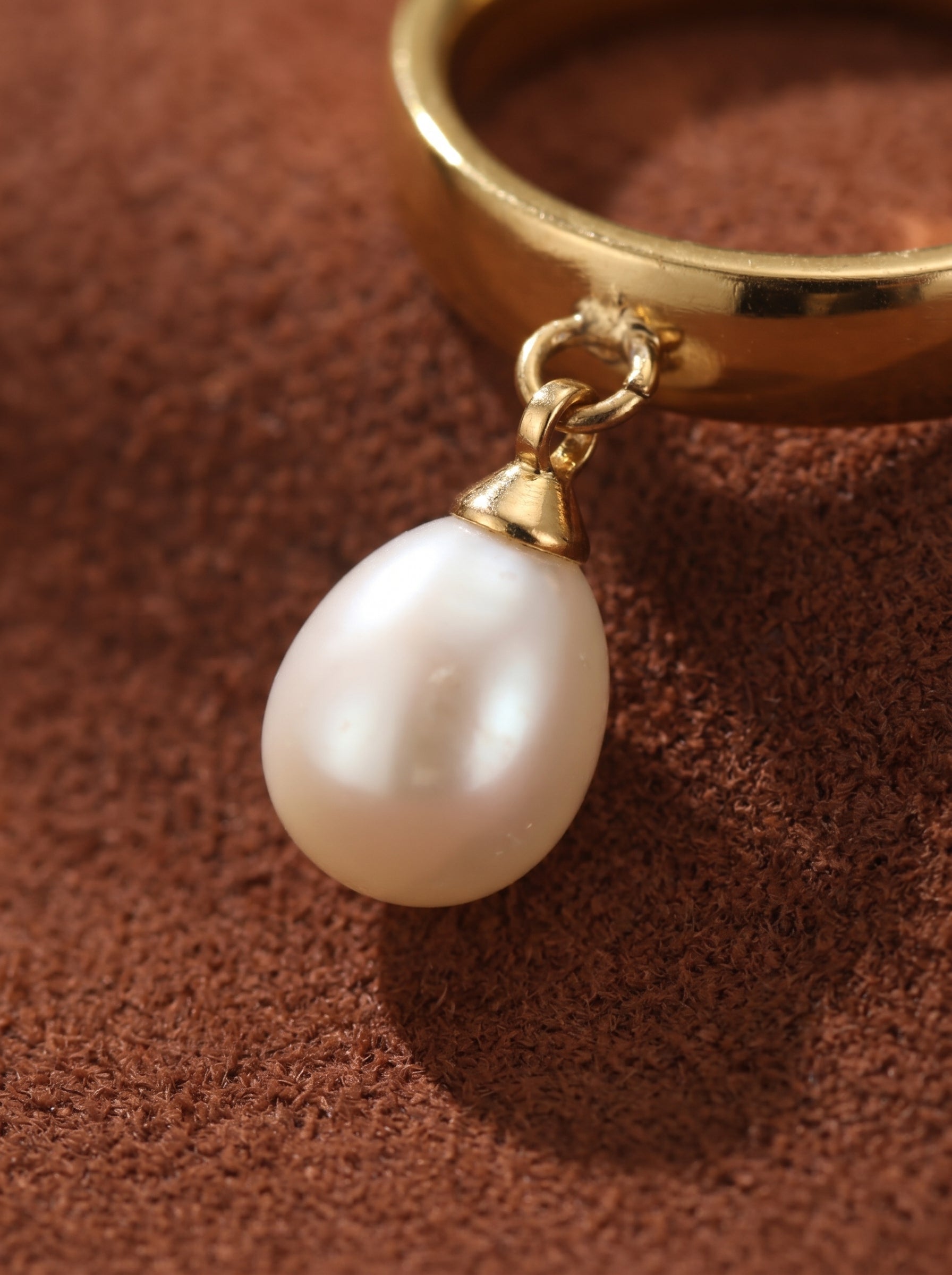 Serene - Gold ring with a dangling white freshwater pearl bead, detailed close-up shot on a brown textured surface.@pearl