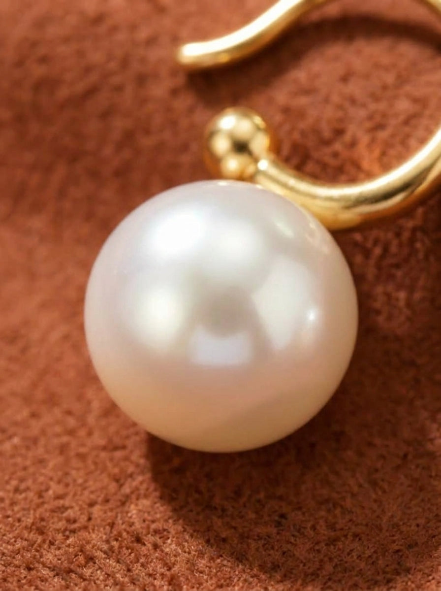 Close-up of a luminous 10mm round freshwater pearl dangling from a sleek 18K gold-plated sterling silver hook, resting on a textured brown surface. @pearl