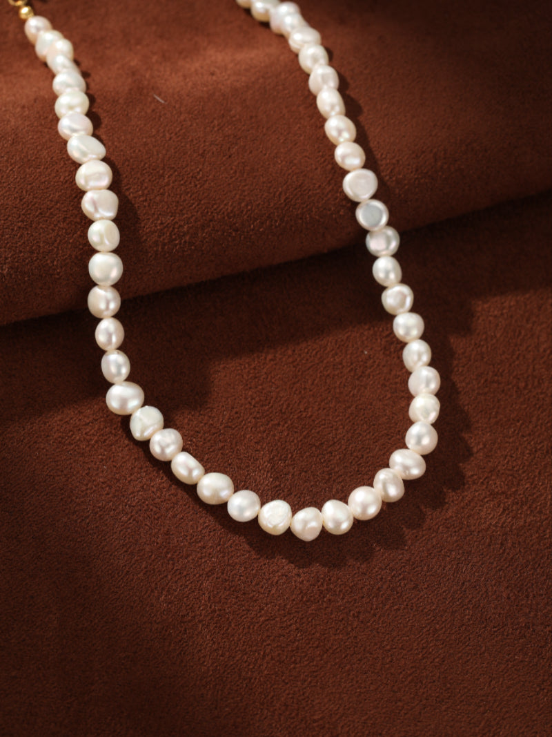 A close-up shot of the Serene Strand Freshwater Pearl Choker Necklace, showcasing its lustrous 6mm white button pearls strung together on a simple gold chain against a brown textured background.