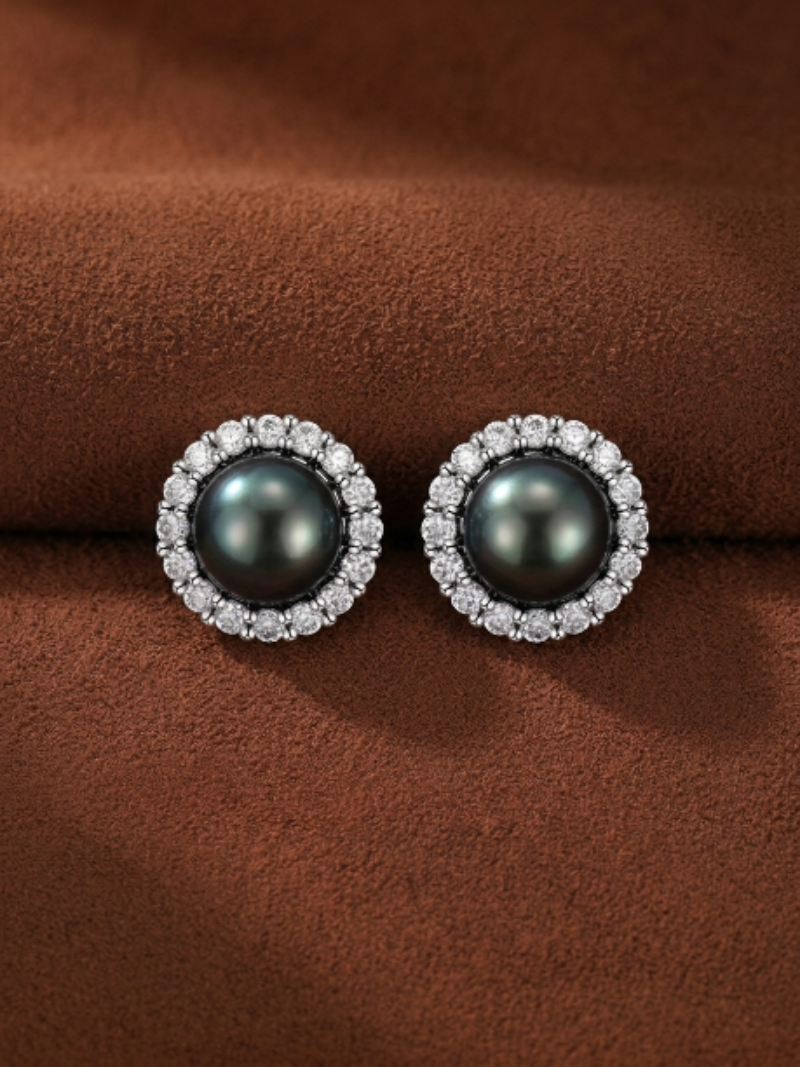 A pair of Shadowlight Tahitian pearl halo stud earrings with a dark, lustrous pearl center surrounded by a halo of sparkling accent stones, set against a textured brown background.