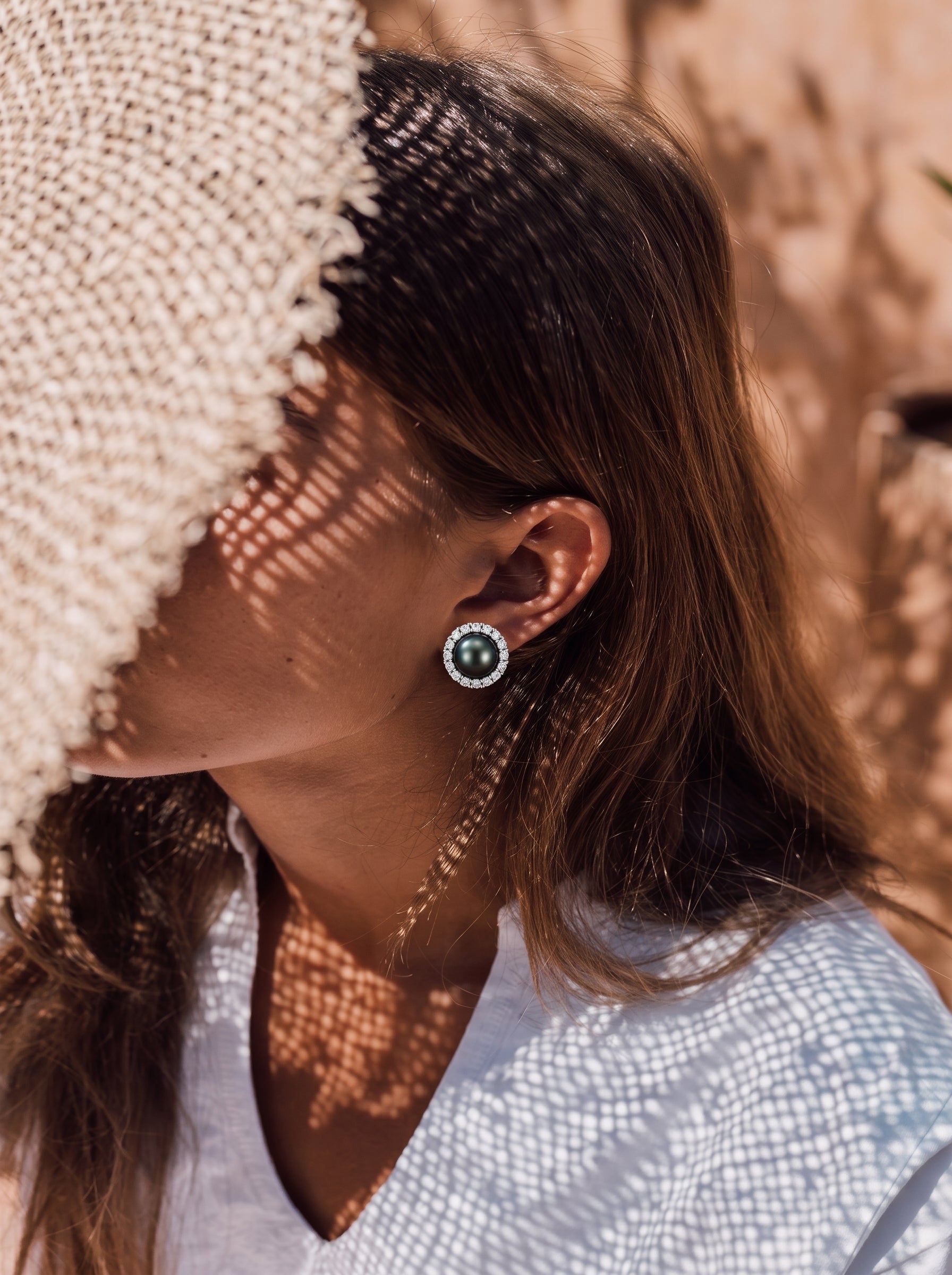 A woman wears Shadowlight Tahitian Pearl Halo Stud Earrings, featuring a dark pearl surrounded by a halo of small diamonds, under a straw hat. The scene suggests a sunny outdoor setting, possibly a vacation. @lifestyle